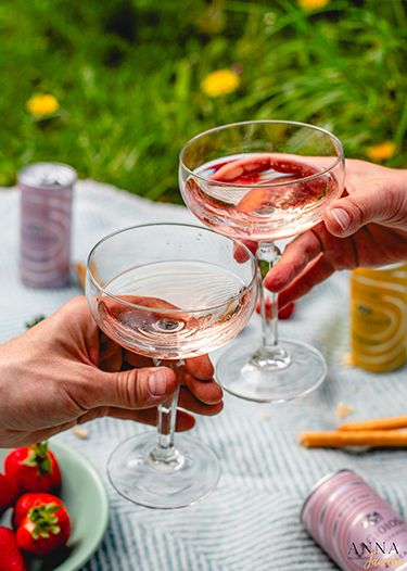 Multiple champagne coupes toasting on a white tablecloth outdoors.