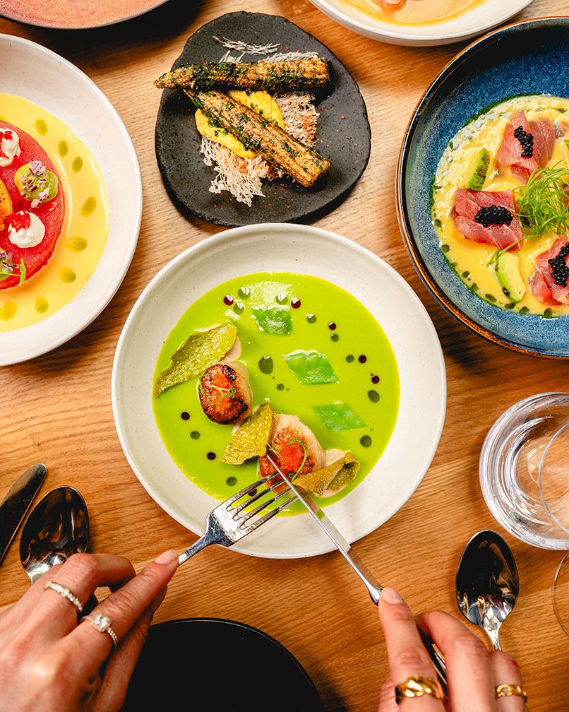 Hands using fork and knife to eat vibrant green soup with figs, surrounded by gourmet dishes on a wooden table.