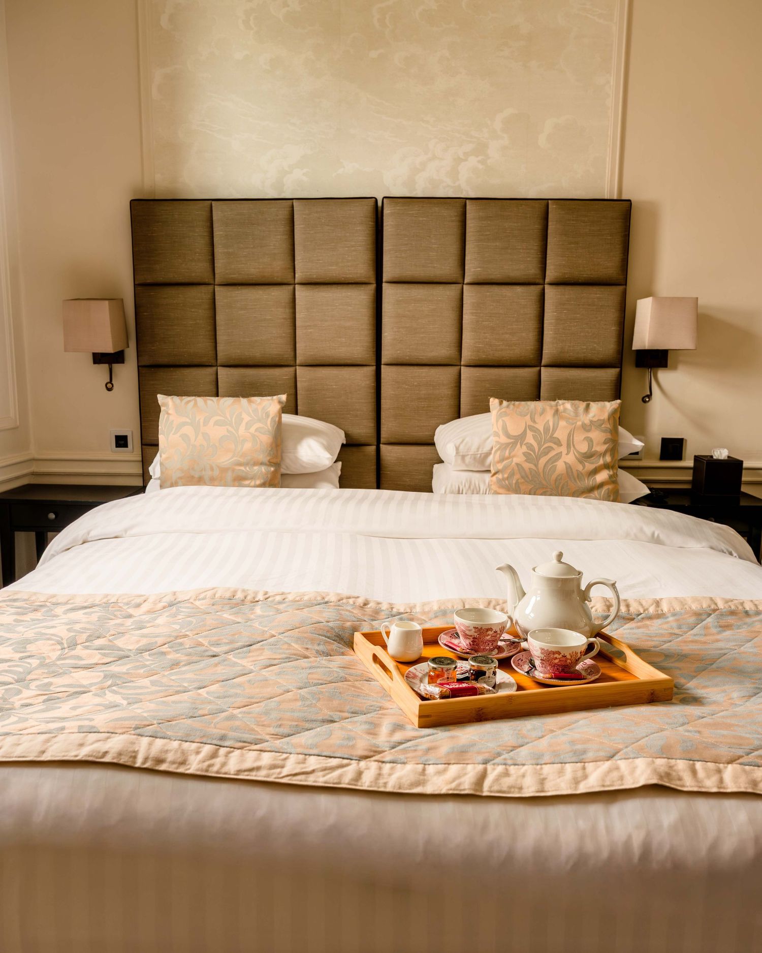 Luxury hotel bedroom with tufted headboard, white bedding, and a wooden breakfast tray with tea set and fruit.