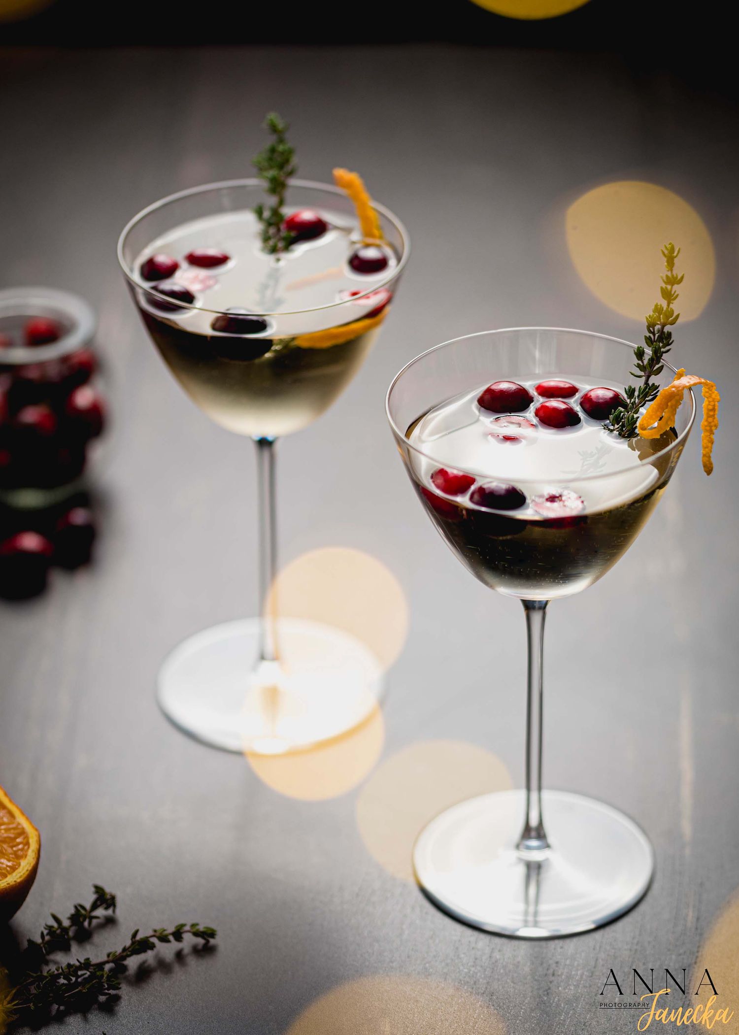 Two cocktails garnished with cranberries and herbs in coupe glasses with bokeh lighting.