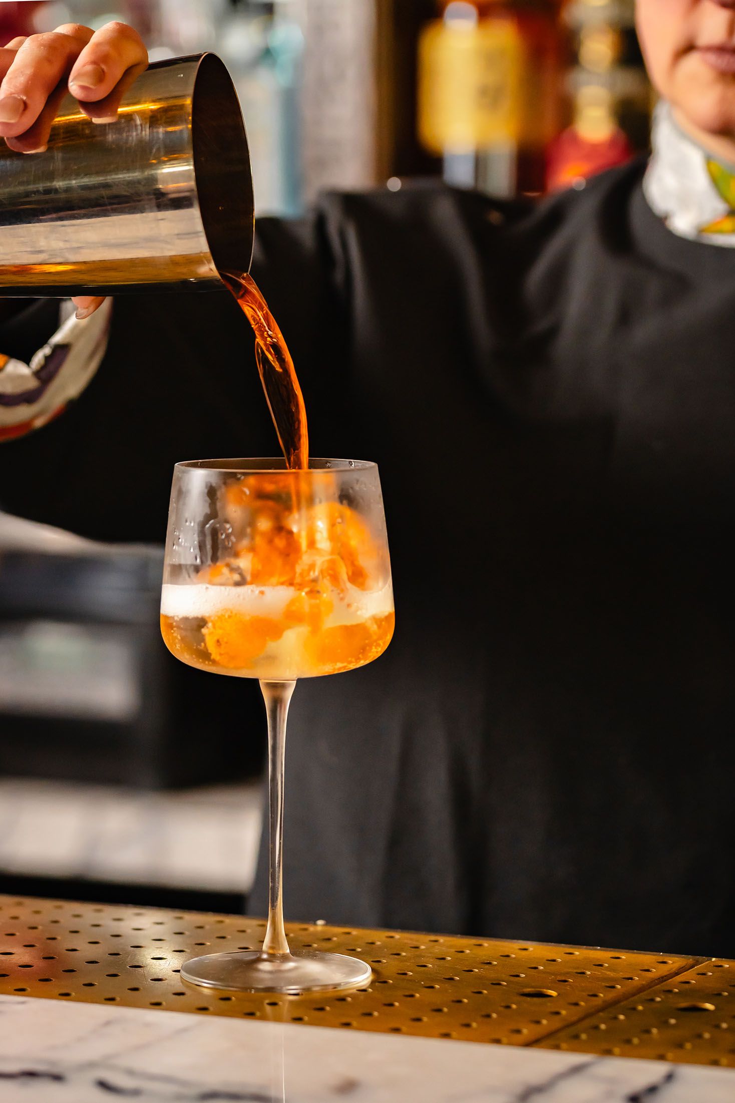 Bartender pouring an orange cocktail from a shaker into a wine glass with ice at a bar.