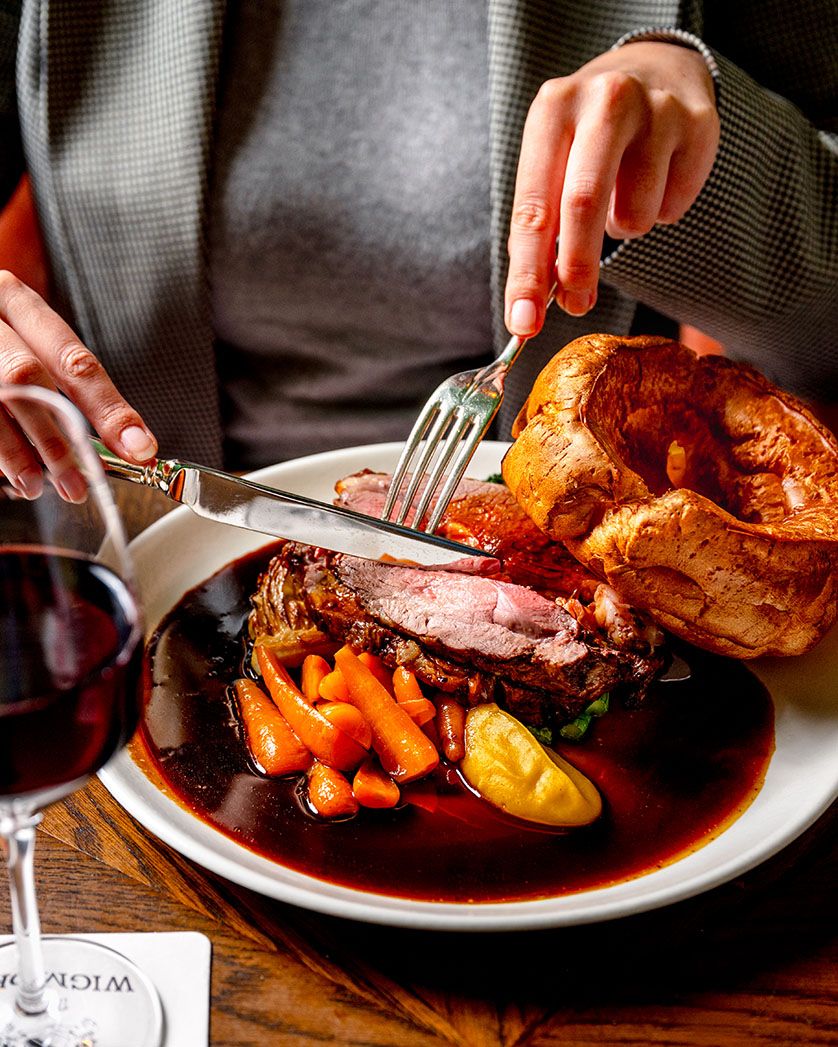Person cutting into a classic British roast beef Sunday dinner with Yorkshire pudding, carrots, and rich gravy on a white plate.