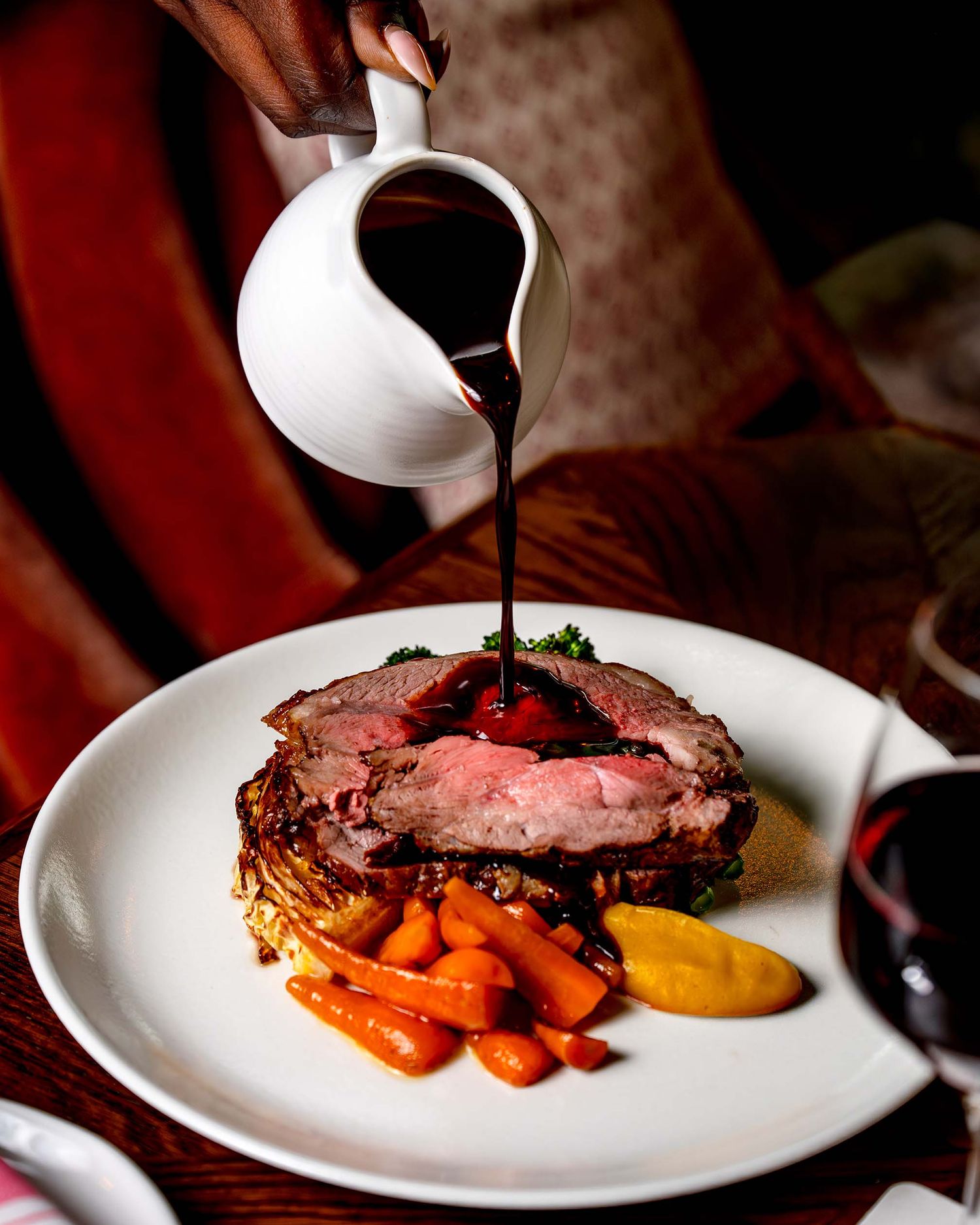 A rich dark sauce being poured over a perfectly cooked prime rib with glazed carrots and mustard on a white plate.