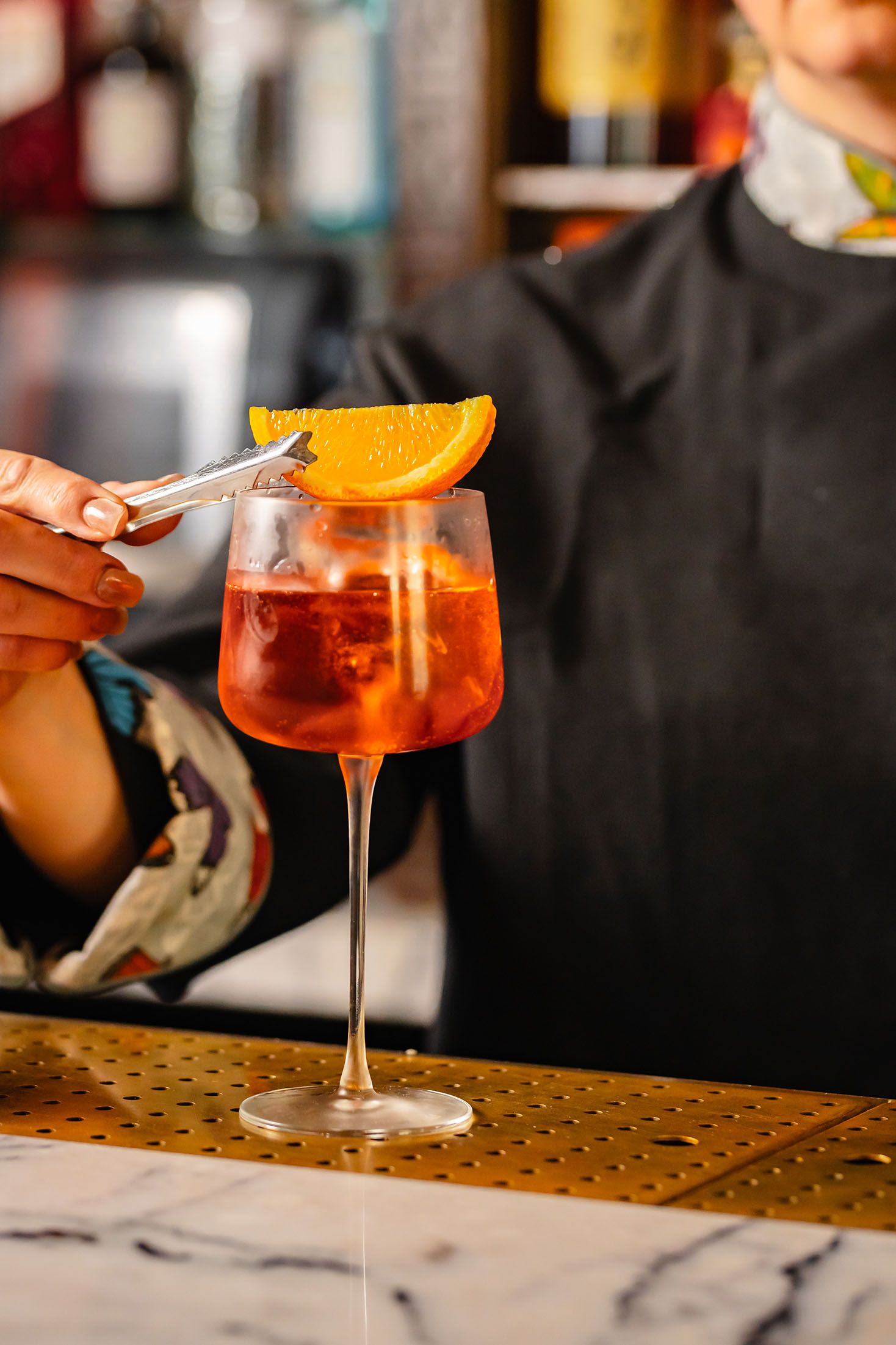 Bartender garnishing a red Aperol Spritz cocktail with an orange slice using tongs at a marble bar.