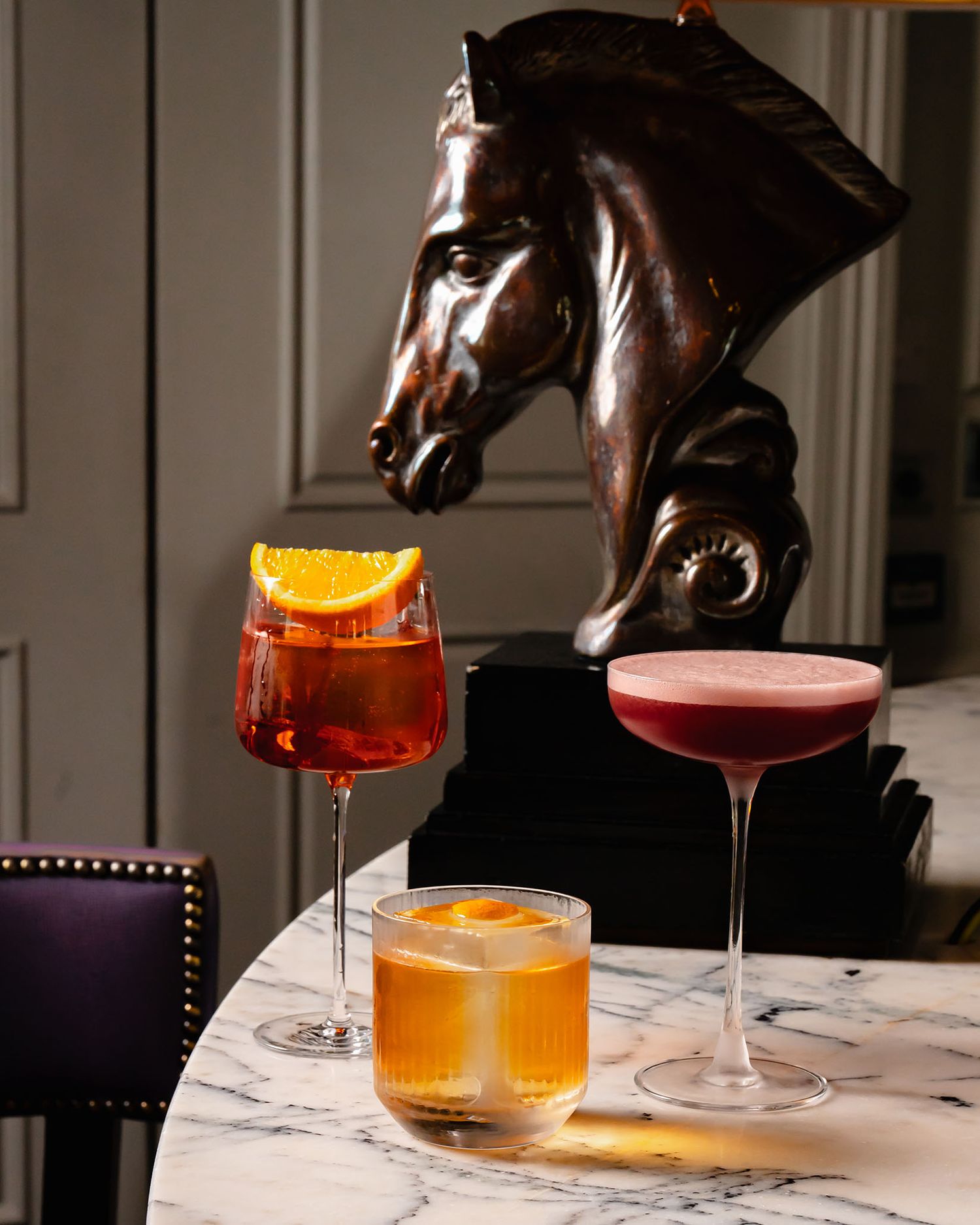 Three elegant cocktails on marble table beside bronze horse sculpture in upscale bar setting.