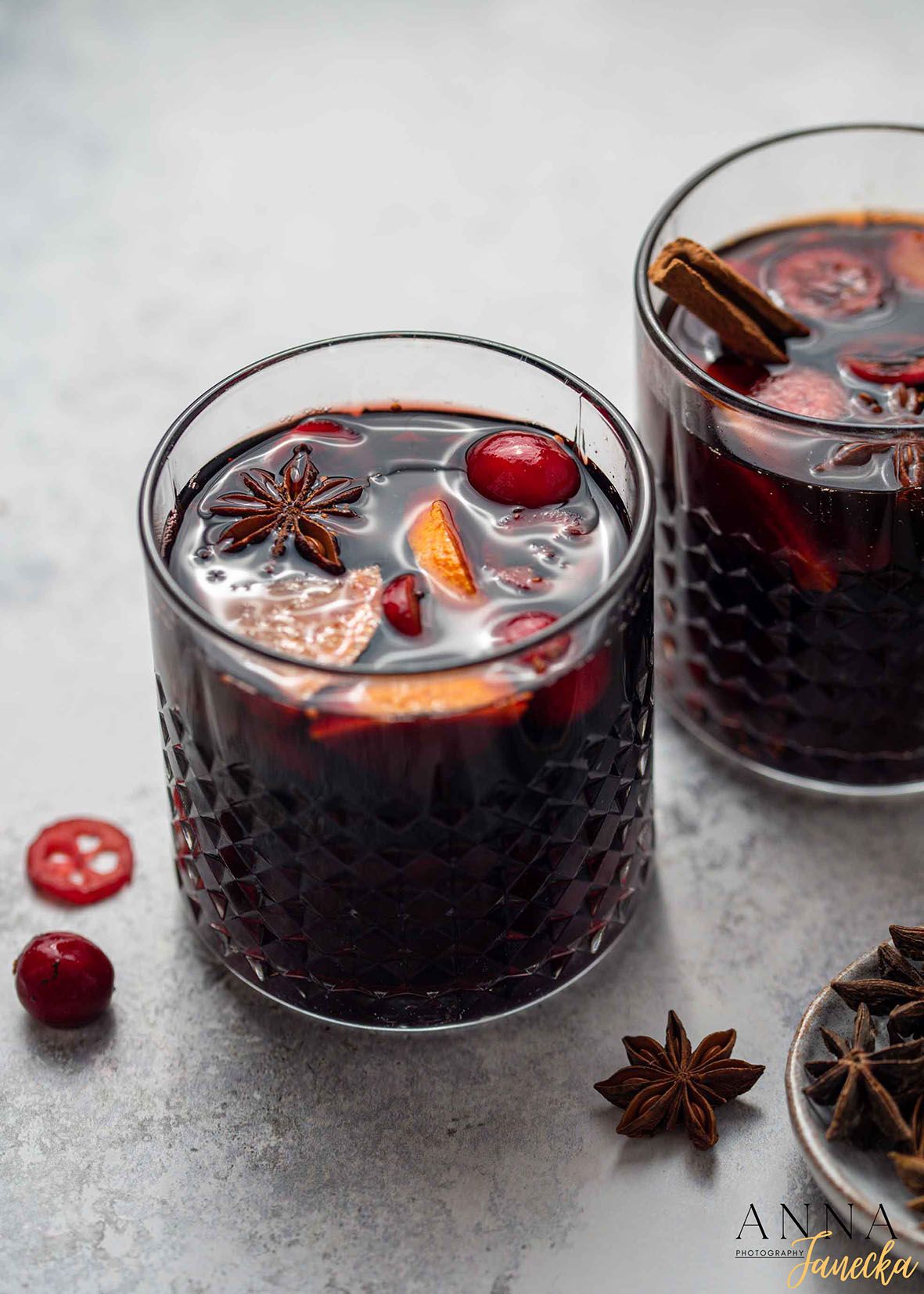 Two dark mulled wine cocktails garnished with star anise, orange slices and cranberries in textured glasses.