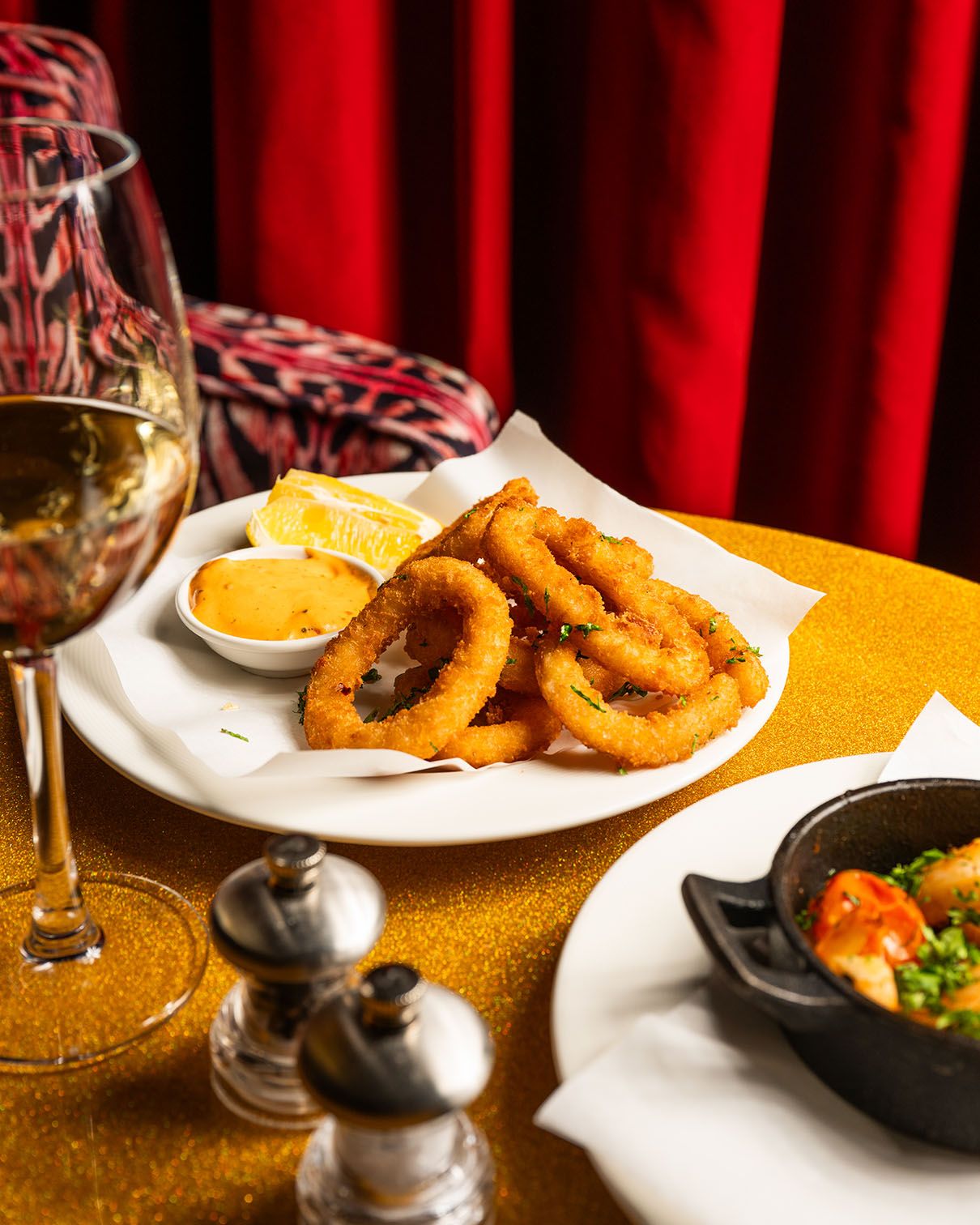 Golden fried calamari rings served with dipping sauce and lemon on a white plate, paired with white wine on a glittery table.