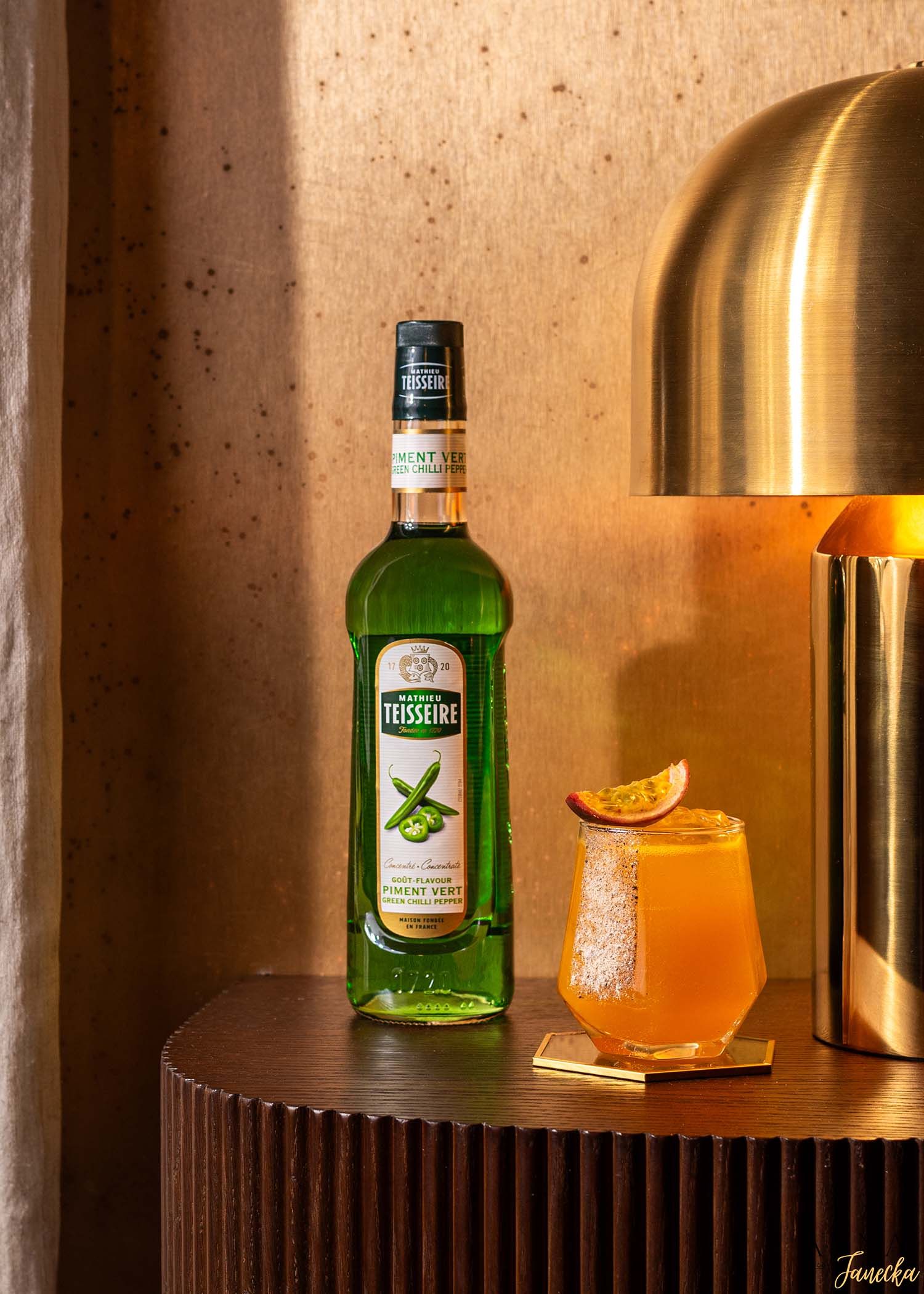 Orange cocktails with citrus garnish displayed with green liquor bottles against warm backlit wall.