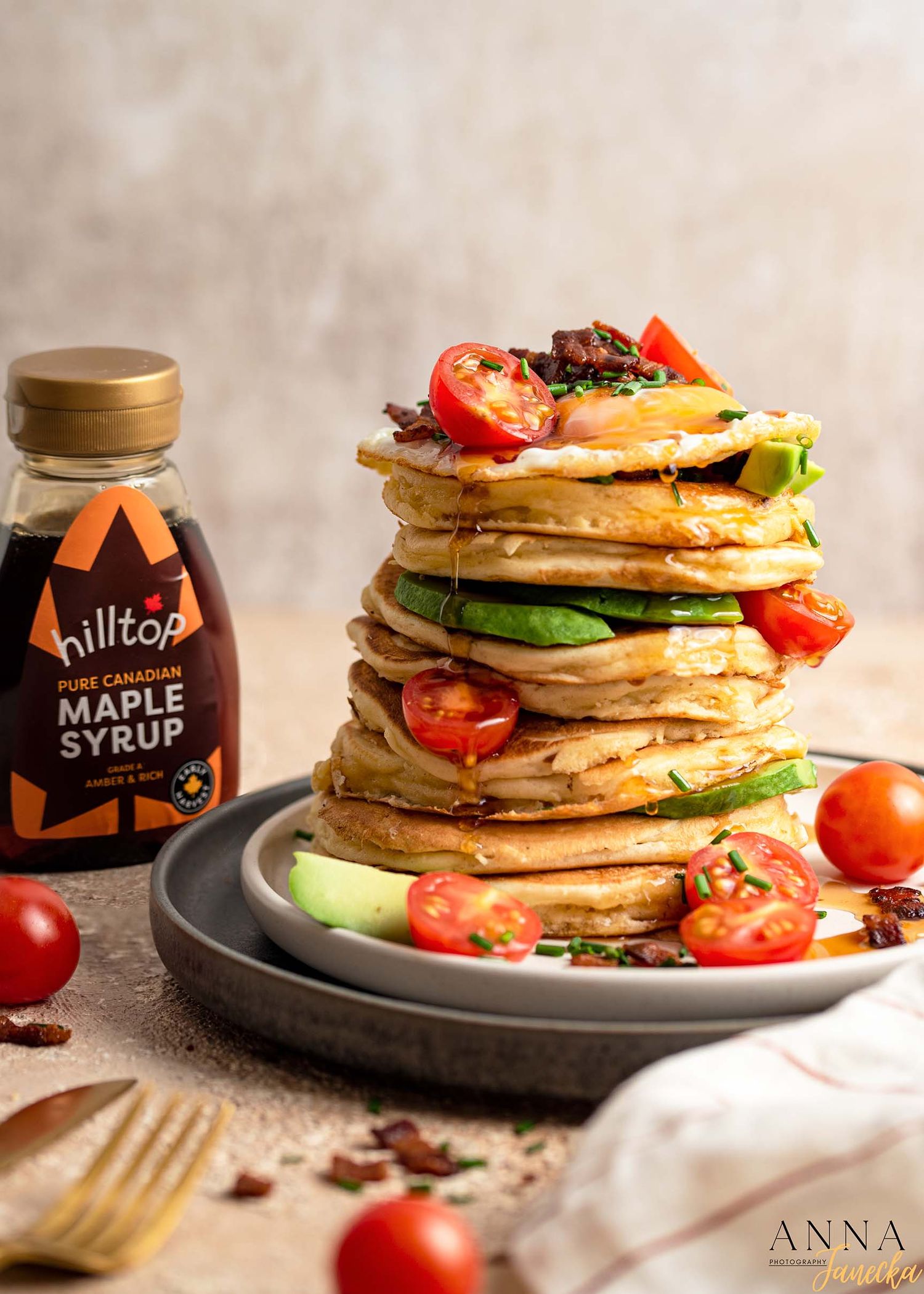 Stack of savory pancakes layered with avocado and tomatoes served with maple syrup.