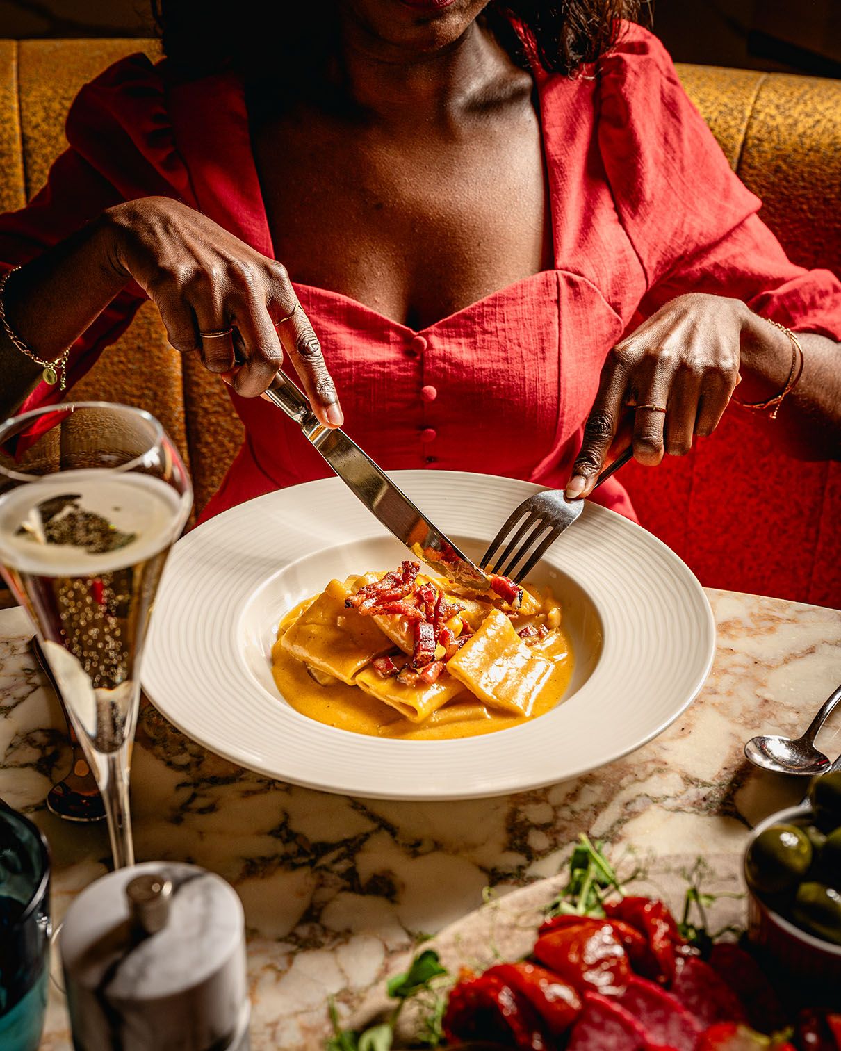 Woman in red dress cutting pasta dish in golden sauce at marble table with champagne glass.