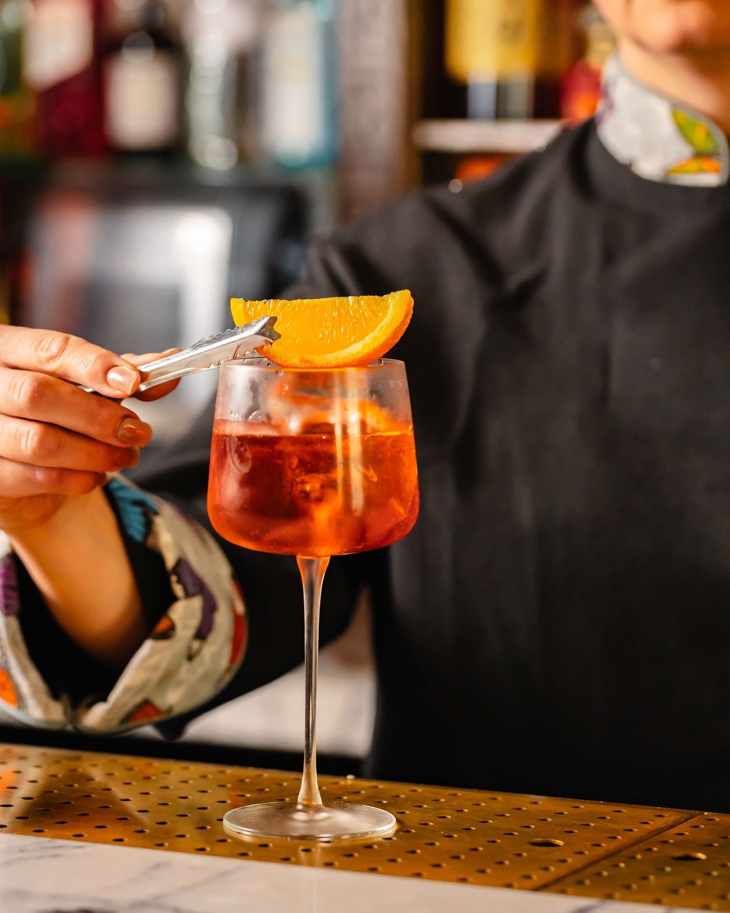 Bartender garnishing a red Aperol Spritz cocktail with an orange slice in an elegant wine glass at a bar.