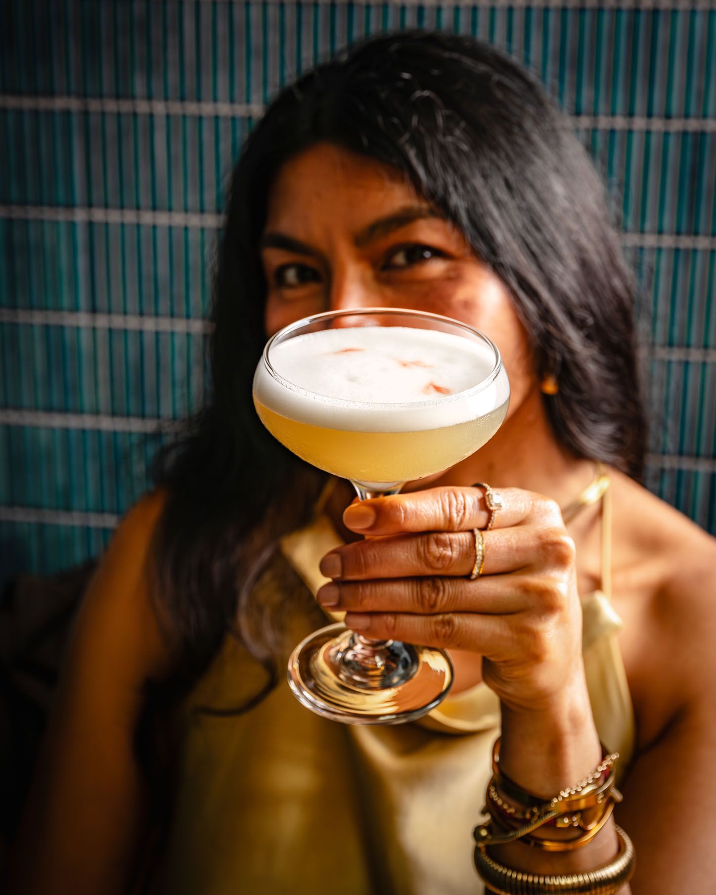 Woman holding a frothy cocktail in a coupe glass, wearing gold jewelry and a gold top, with a teal backdrop.