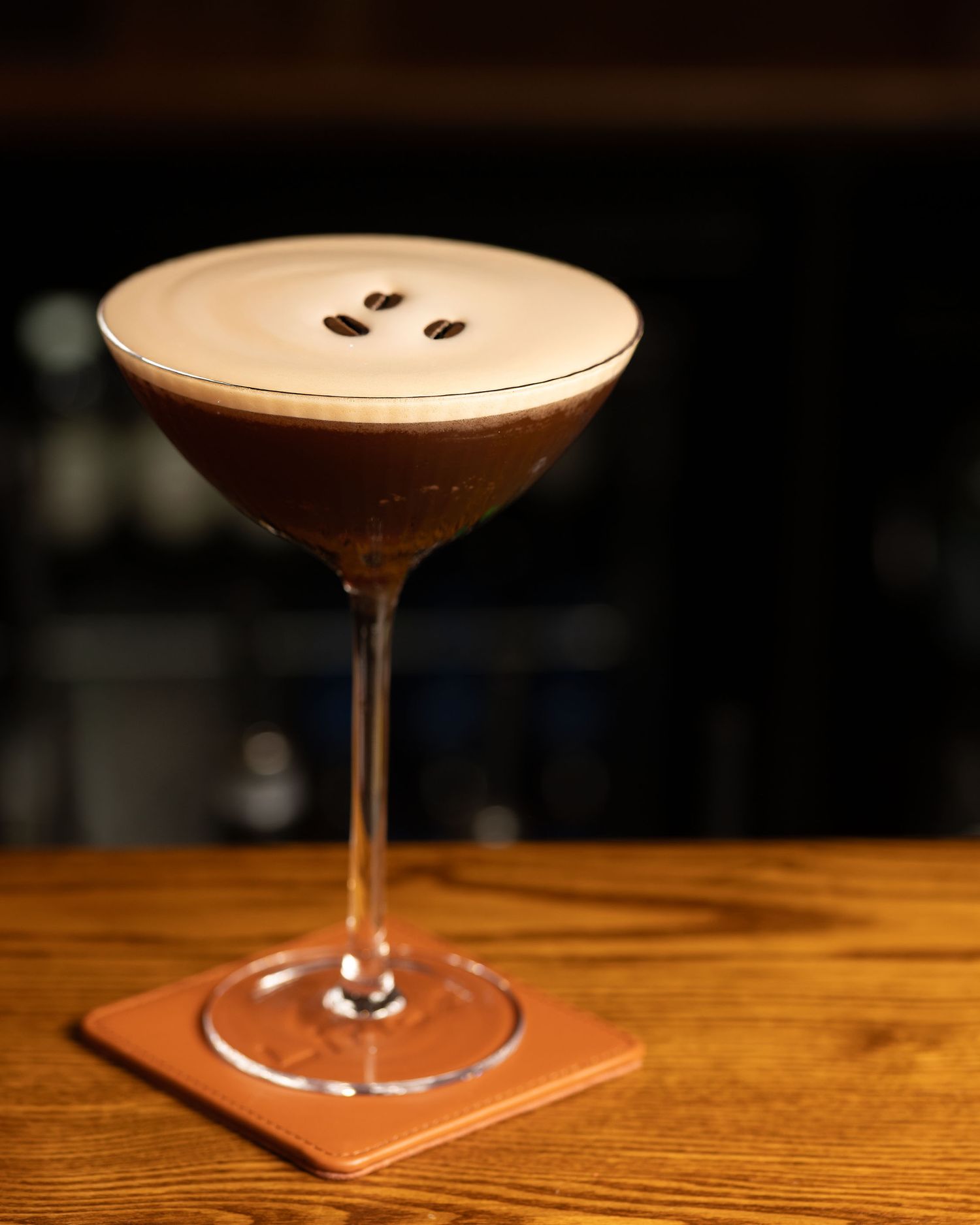 A classic espresso martini in a coupe glass with frothy top and three coffee beans, served on a leather coaster.