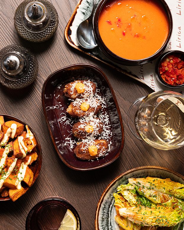 Overhead view of Mediterranean tapas including tomato soup, roasted vegetables, and grilled bread on a dark wooden table.