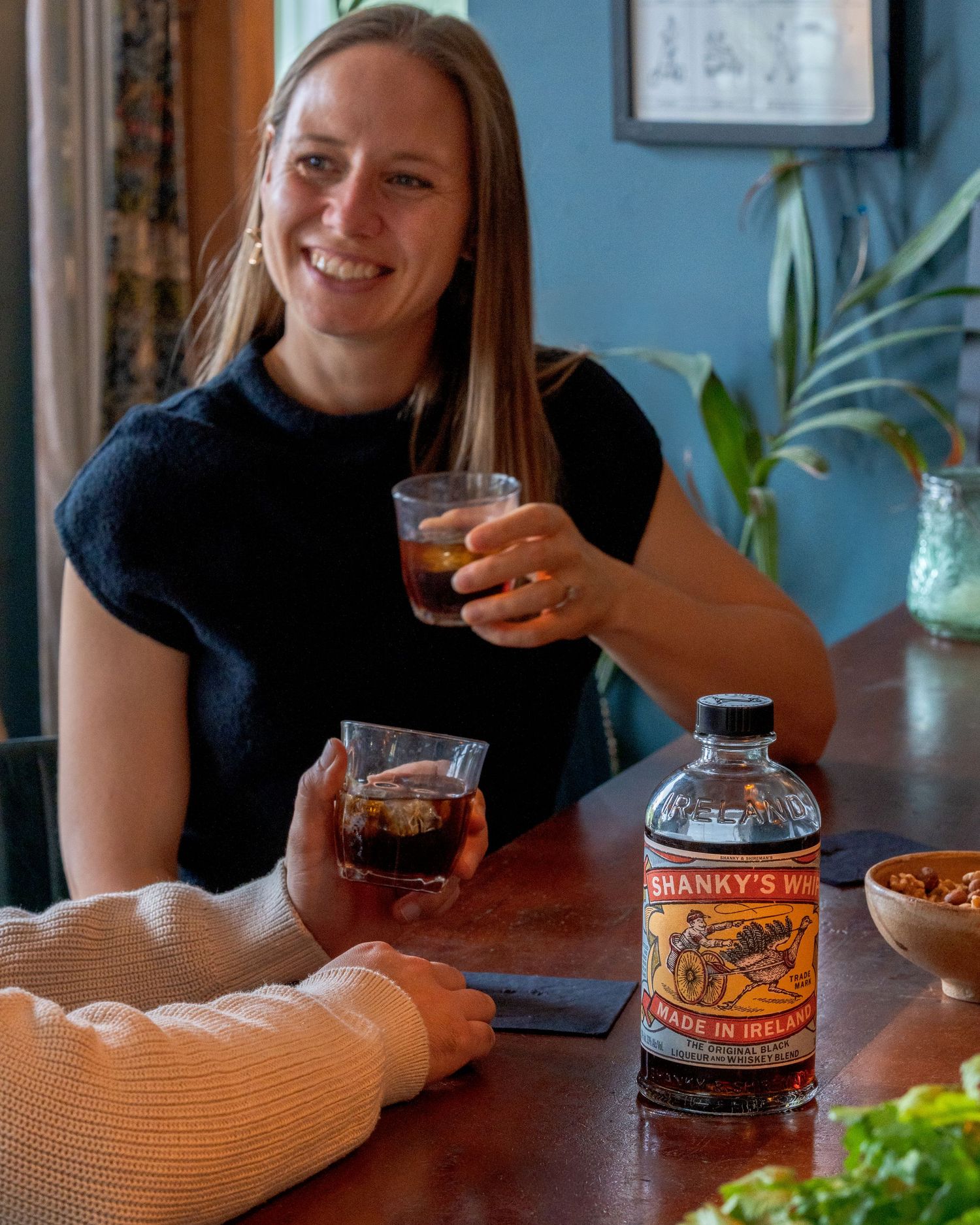 Someone enjoys a cocktail at a cozy table with Hanover's Spirits bottle and snacks in a blue-walled room.