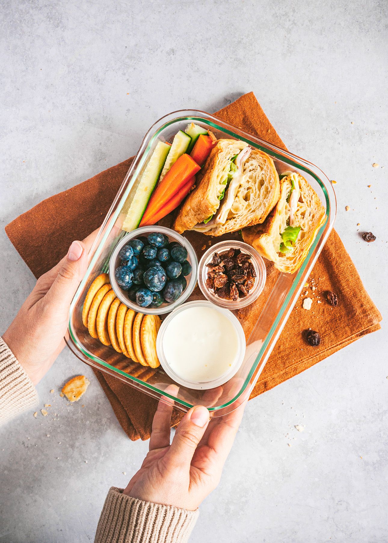Hand arranges healthy snack box with fresh berries, crackers, dip, and dried fruits on a light gray surface.