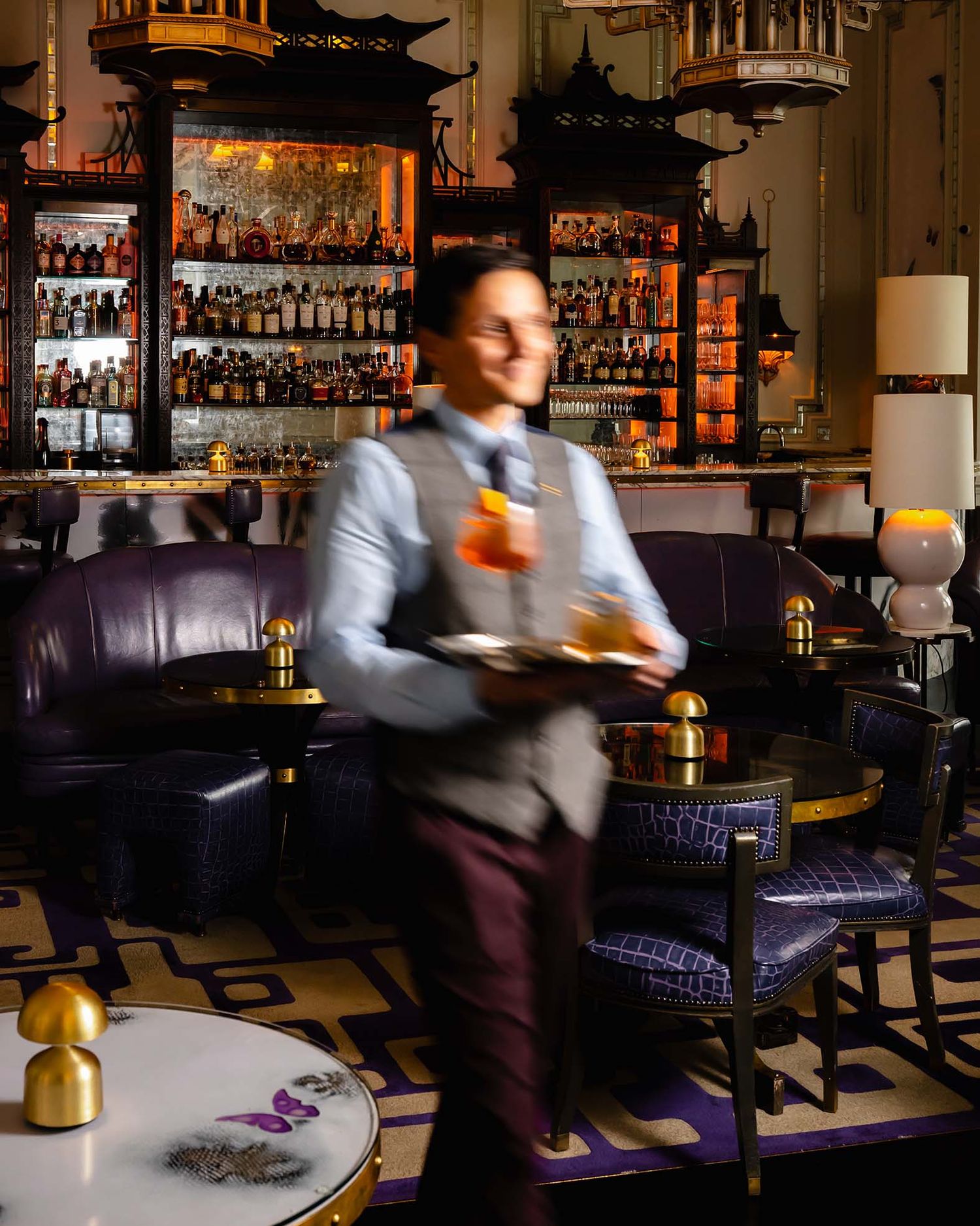 Blurred waiter carrying cocktails through an elegant bar with purple velvet seating and gold decor.