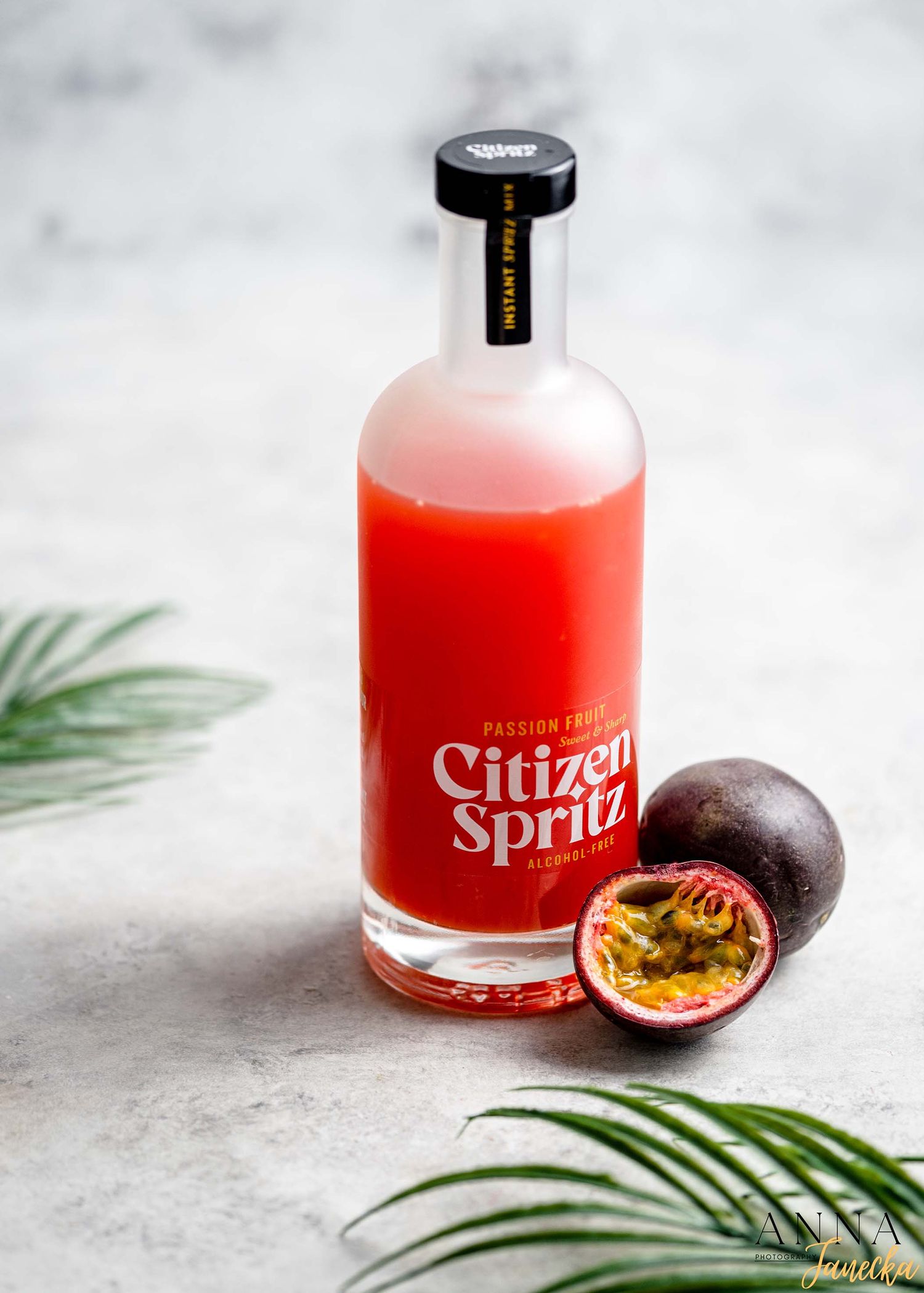 Red citrus cocktail in glass bottles with fresh passion fruit and palm leaves on marble surface.