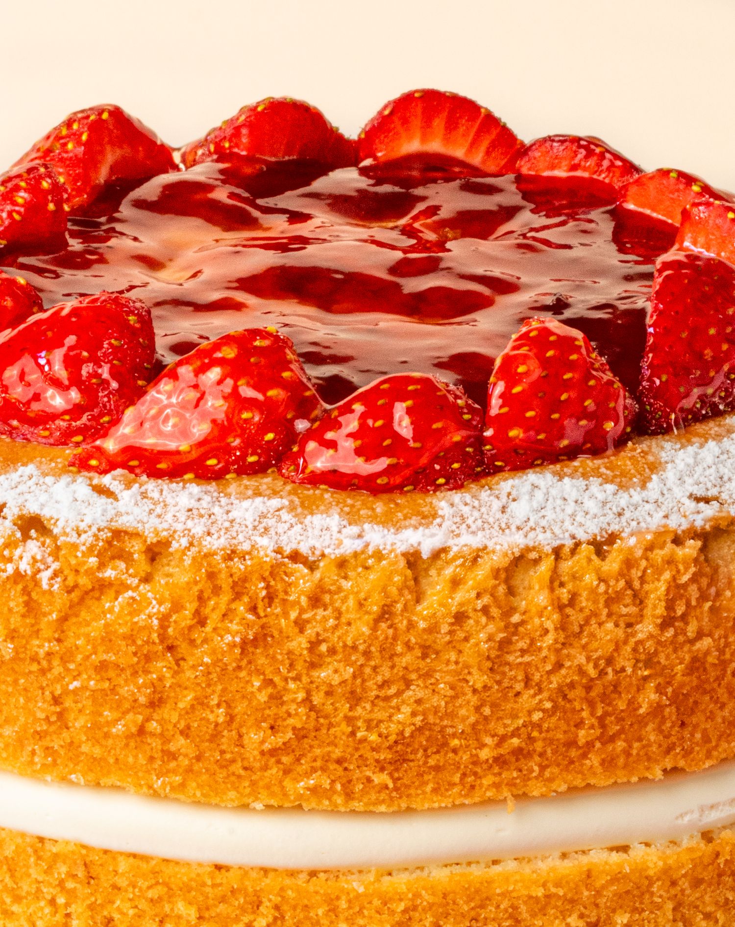 A layered sponge cake topped with fresh strawberries and red glaze, dusted with powdered sugar on a white plate.