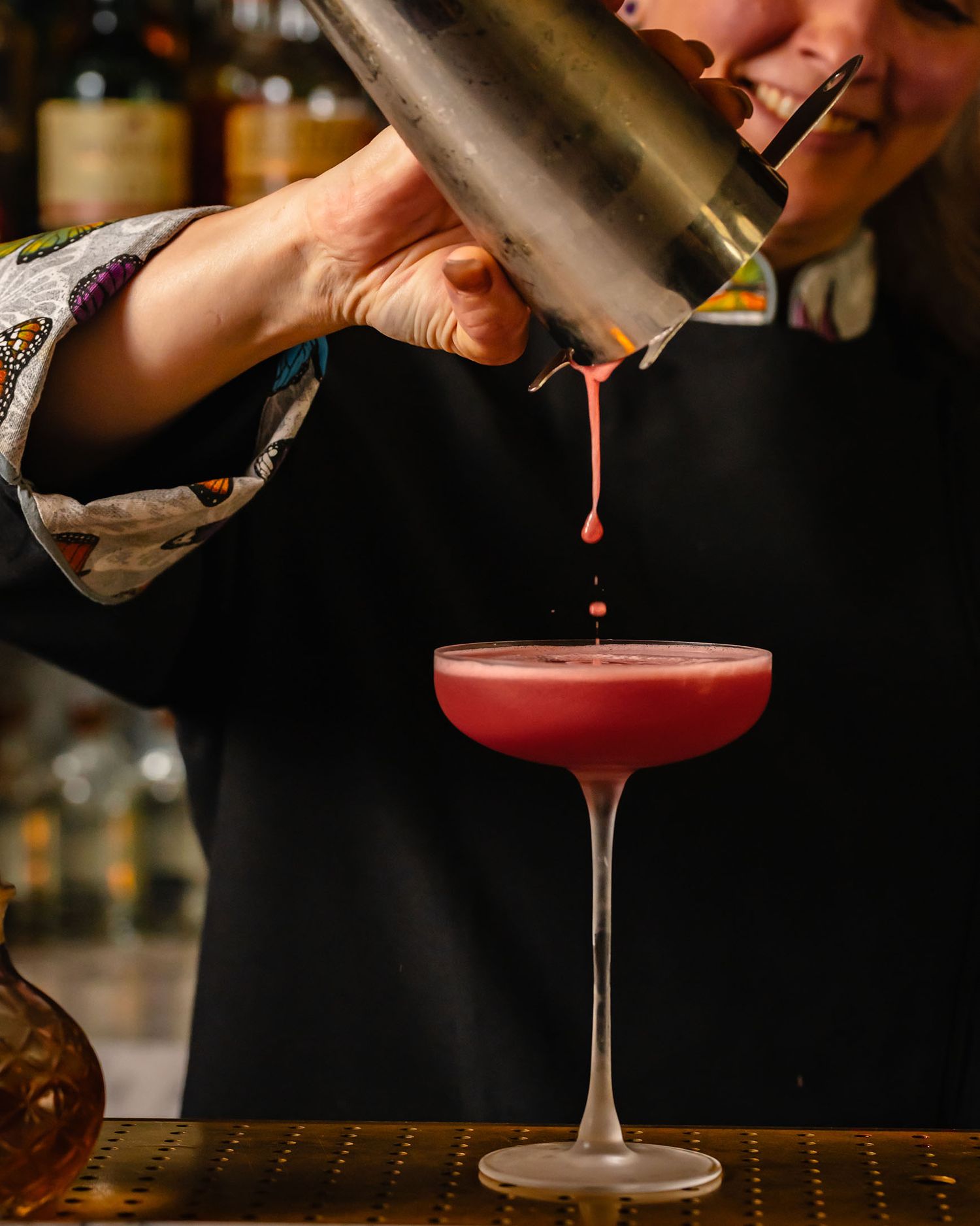 Bartender pouring a pink cocktail into a coupe glass from a shaker at a dimly lit bar.