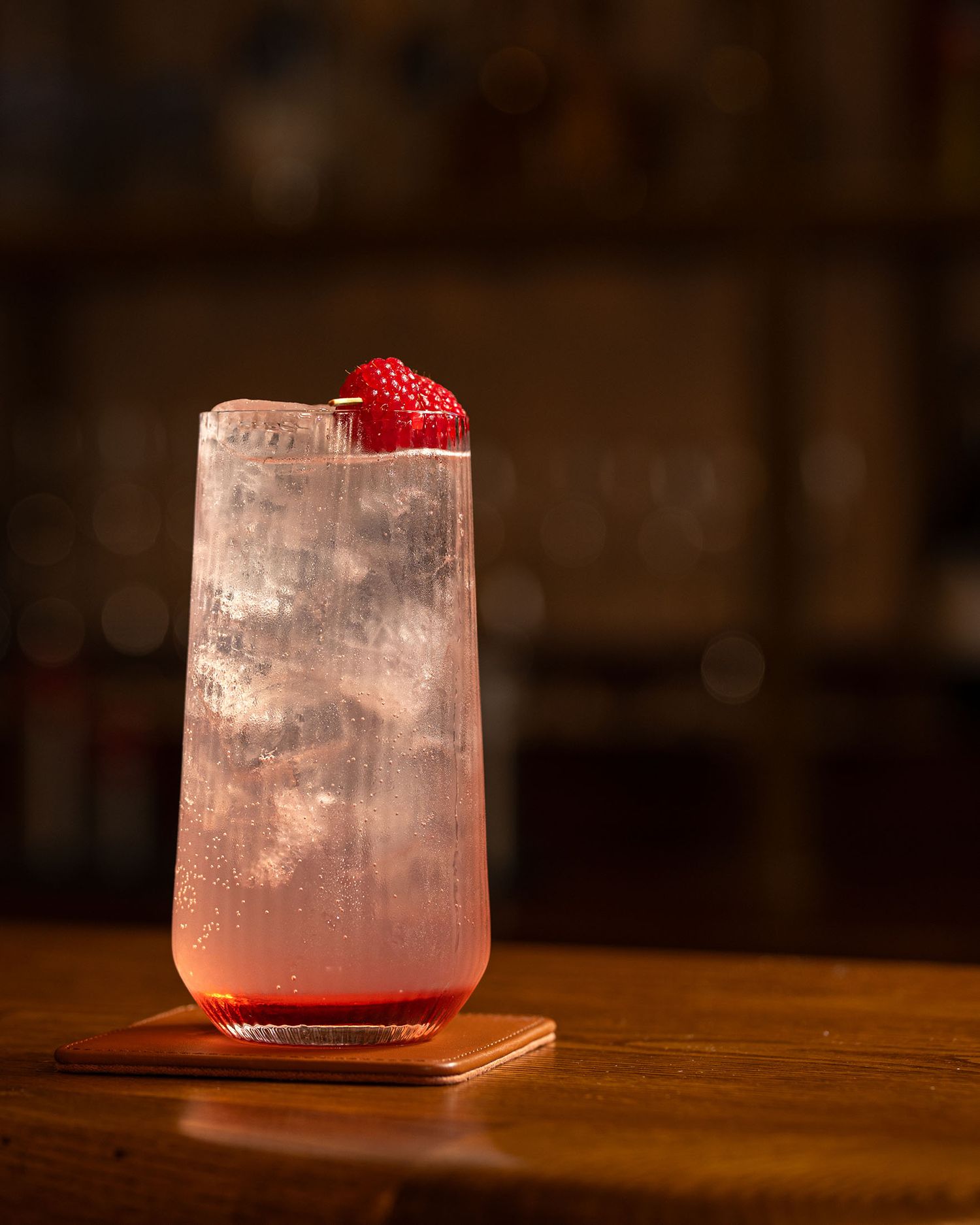 A refreshing pink raspberry cocktail in a tall glass with ice, garnished with fresh raspberries on a wooden bar.