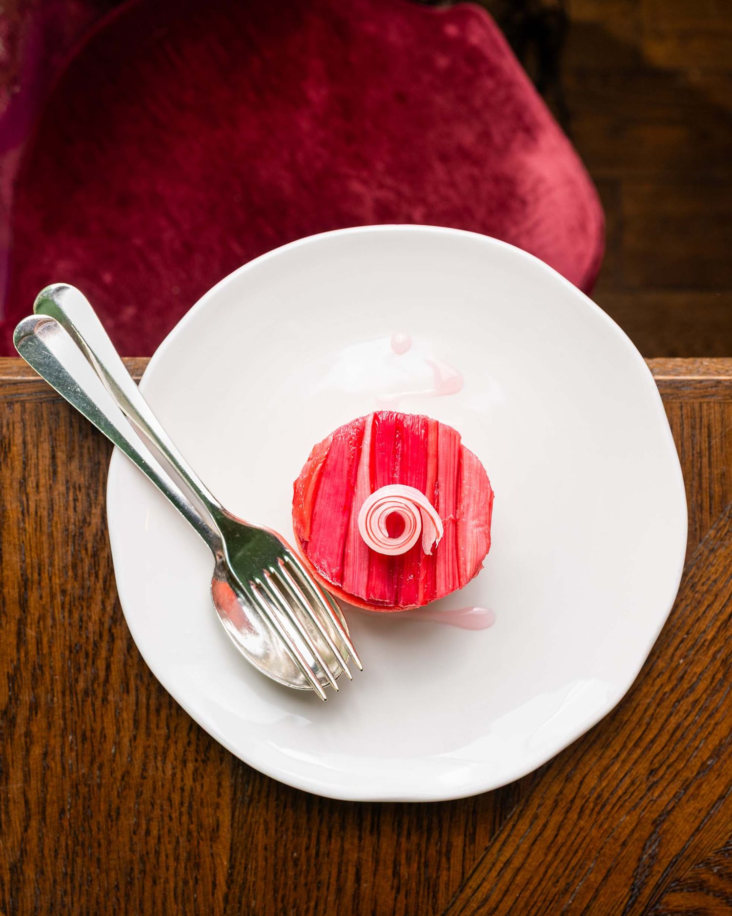 A single pink frosted cupcake with a swirl decoration on a white plate with a silver fork on a wooden table.