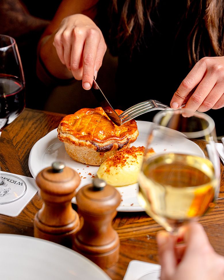 Person cutting into a golden pastry pie on a white plate, served with mash, alongside white and red wine glasses.
