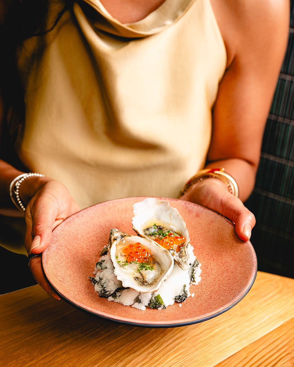 Woman holding a pink plate with fresh oysters topped with caviar and herbs at an upscale restaurant.