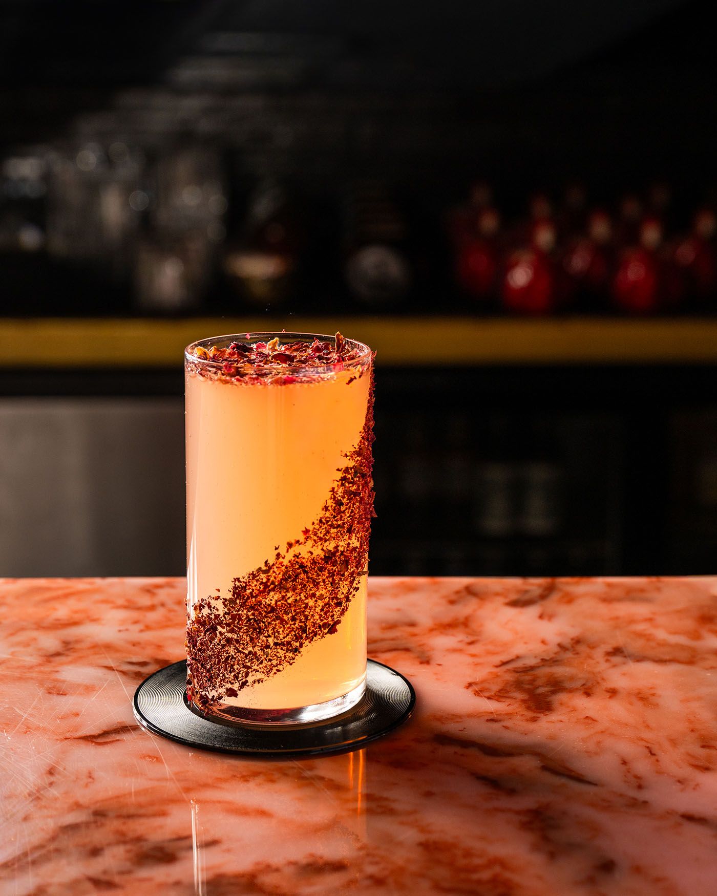 A tall cocktail glass with a pink drink and red spice swirl sits on a marble bar in a dimly lit lounge.