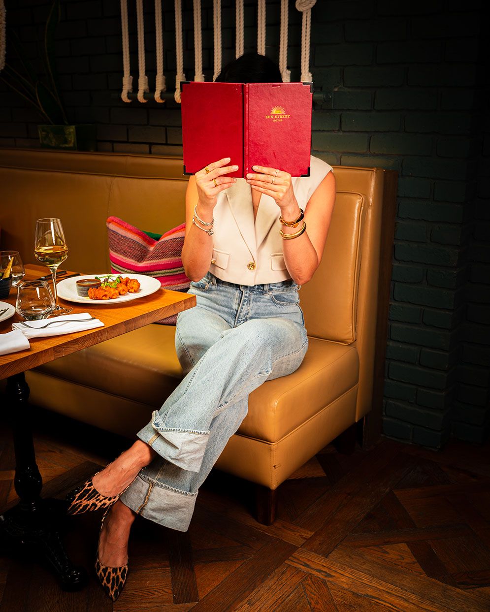 Woman in jeans holding red menu at upscale restaurant booth with wine and food on wooden table.