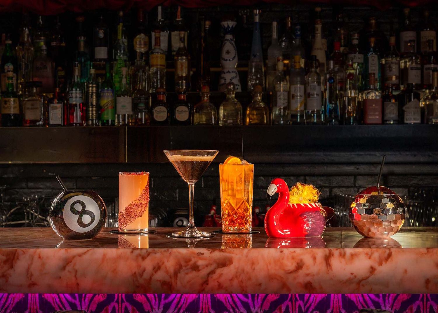 Five colorful cocktails lined up on a marble bar with a backlit liquor shelf in a dimly lit lounge.
