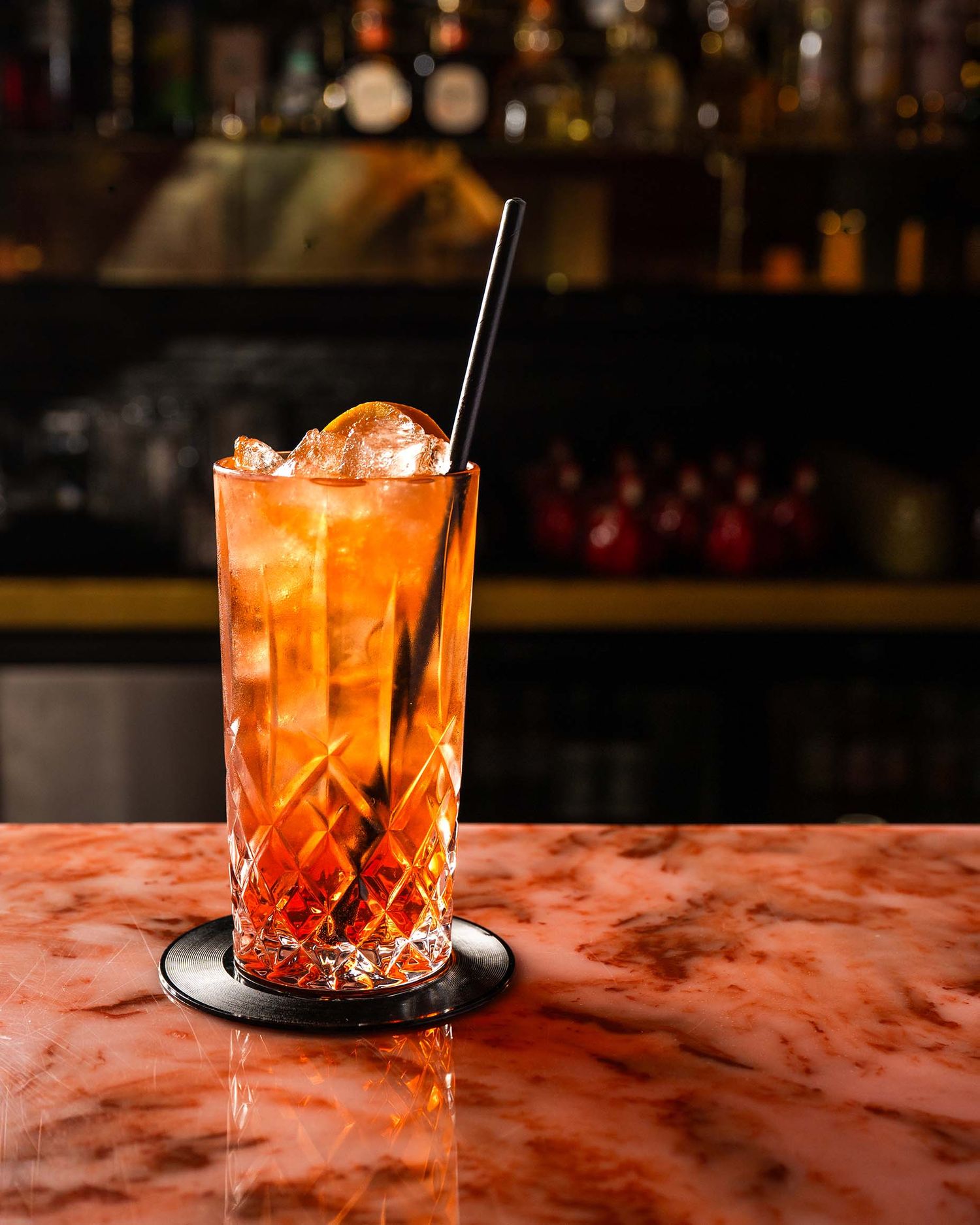 A vibrant amber cocktail in a crystal highball glass with a black straw, served on a marble bar top.