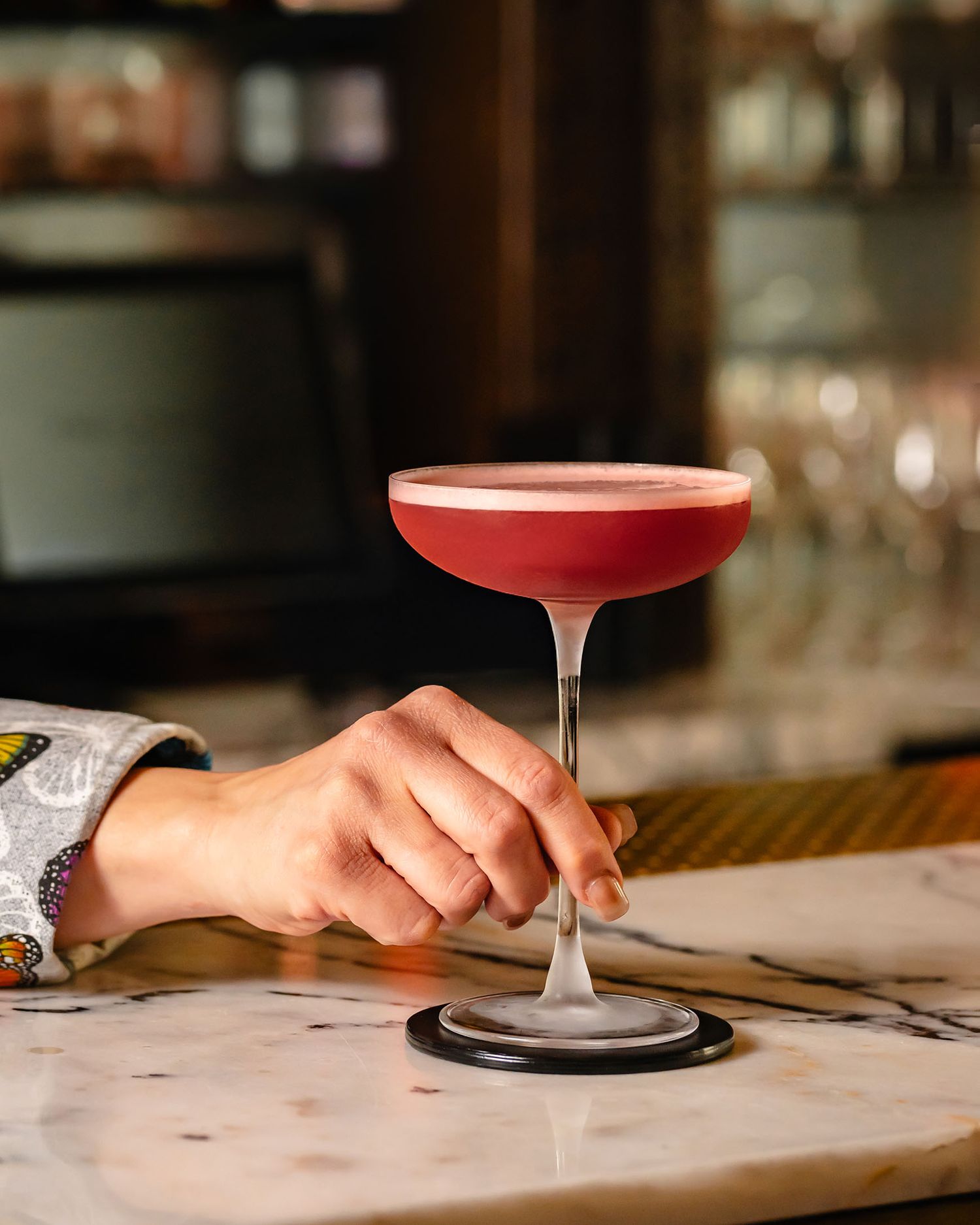 Hand holding a pink cocktail in a coupe glass on a marble bar top in a dimly lit upscale cocktail lounge.