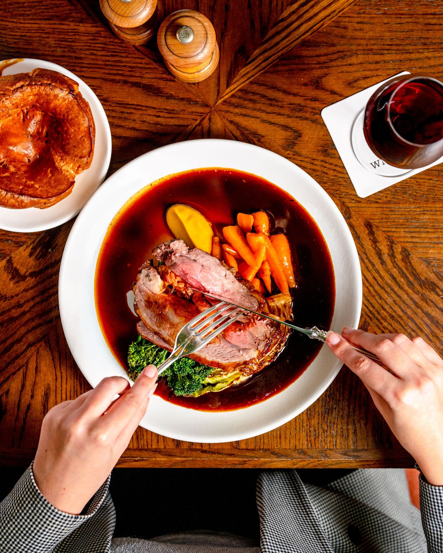 Overhead view of sliced duck breast with vegetables in red wine sauce, hands cutting meat with knife and fork on wooden table.