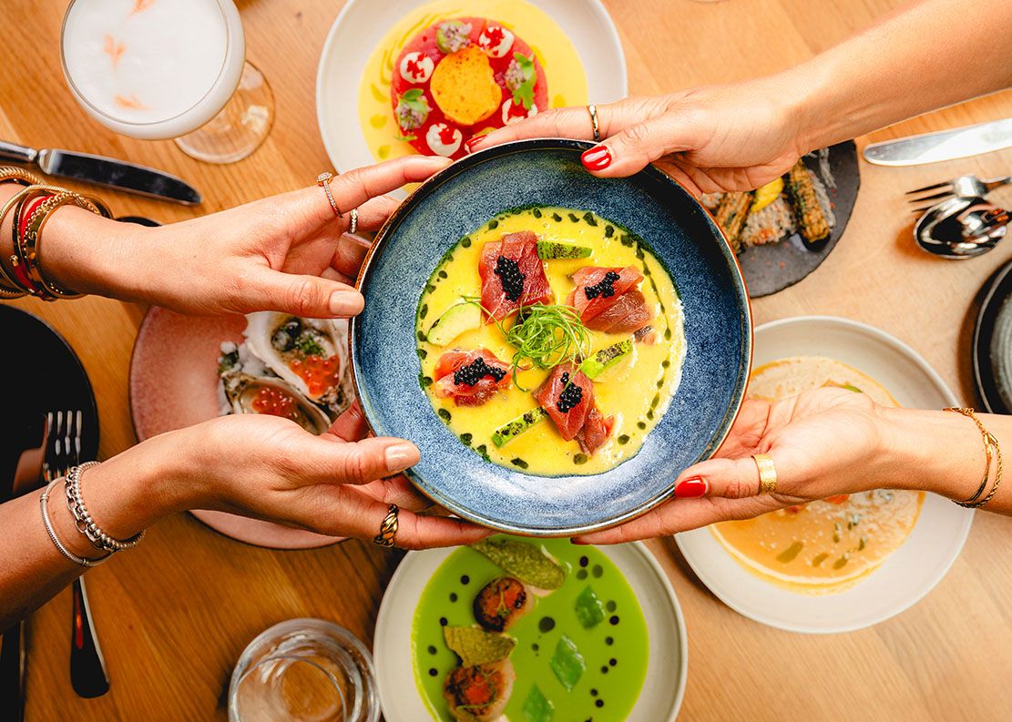 Friends sharing vibrant gourmet dishes around a wooden table, hands reaching for a colorful seafood plate.