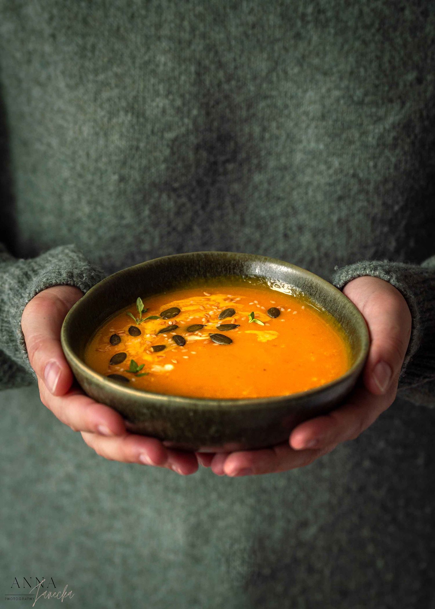 Hands holding bowl of creamy pumpkin soup garnished with seeds against dark background.