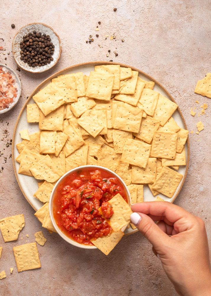 A series of hands reaching for tortilla chips arranged around a bowl of red salsa on a gray textured surface.