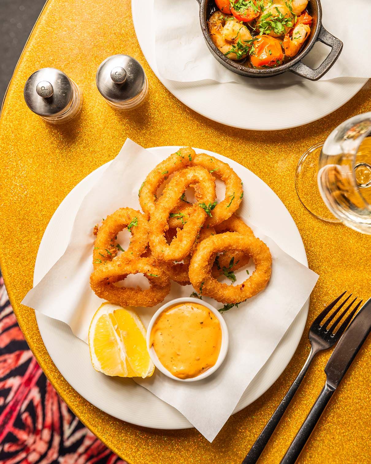 Golden fried calamari rings served with lemon wedges and dipping sauce on a white plate atop a yellow tablecloth.
