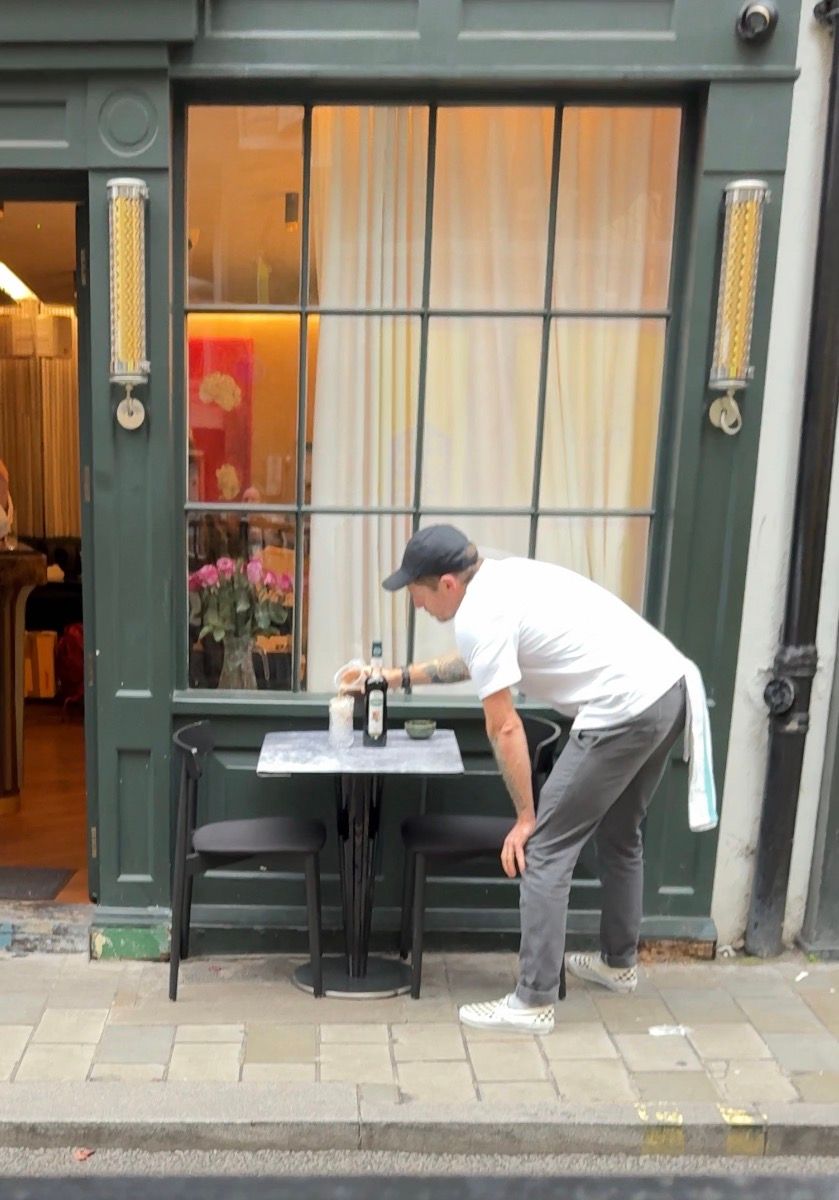 Restaurant staff member cleaning and setting up an outdoor table near a storefront with green trim and large windows.