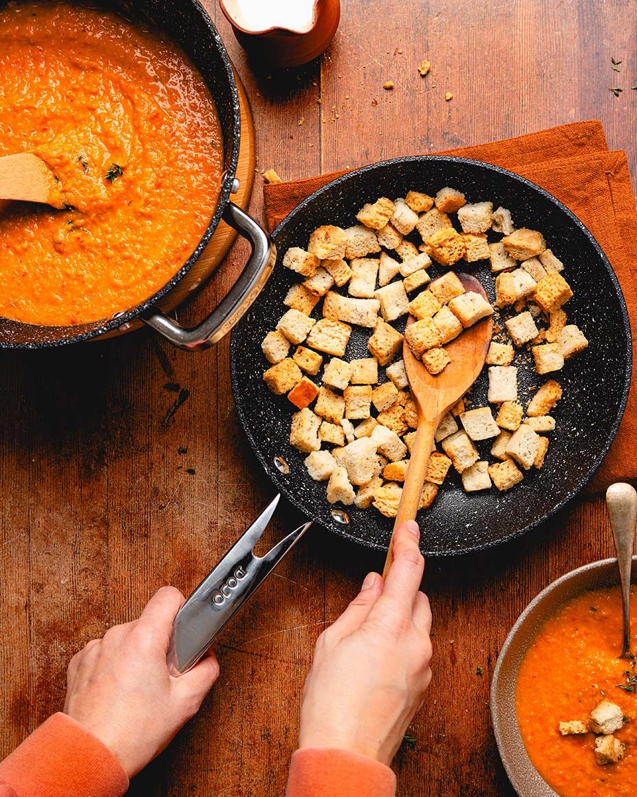 Hands stirring golden croutons in a skillet beside a pot of creamy tomato soup on a wooden surface.