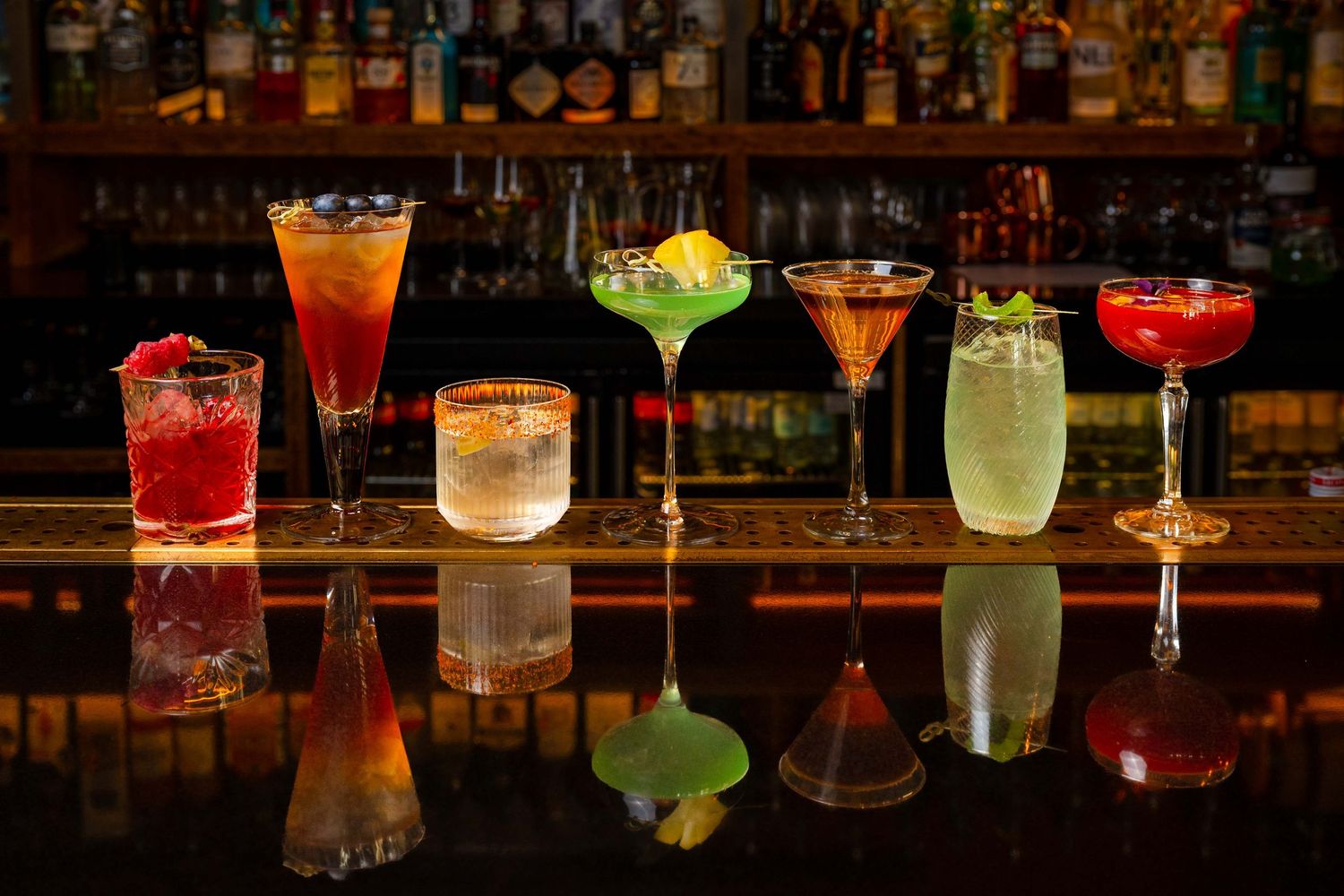 Seven colorful cocktails lined up on a reflective bar counter with a backlit liquor display in the background.