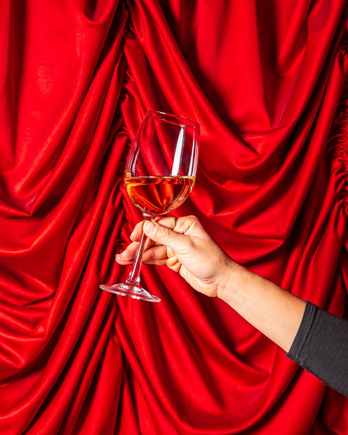 A hand holding a glass of rosé wine against a luxurious red draped fabric backdrop.