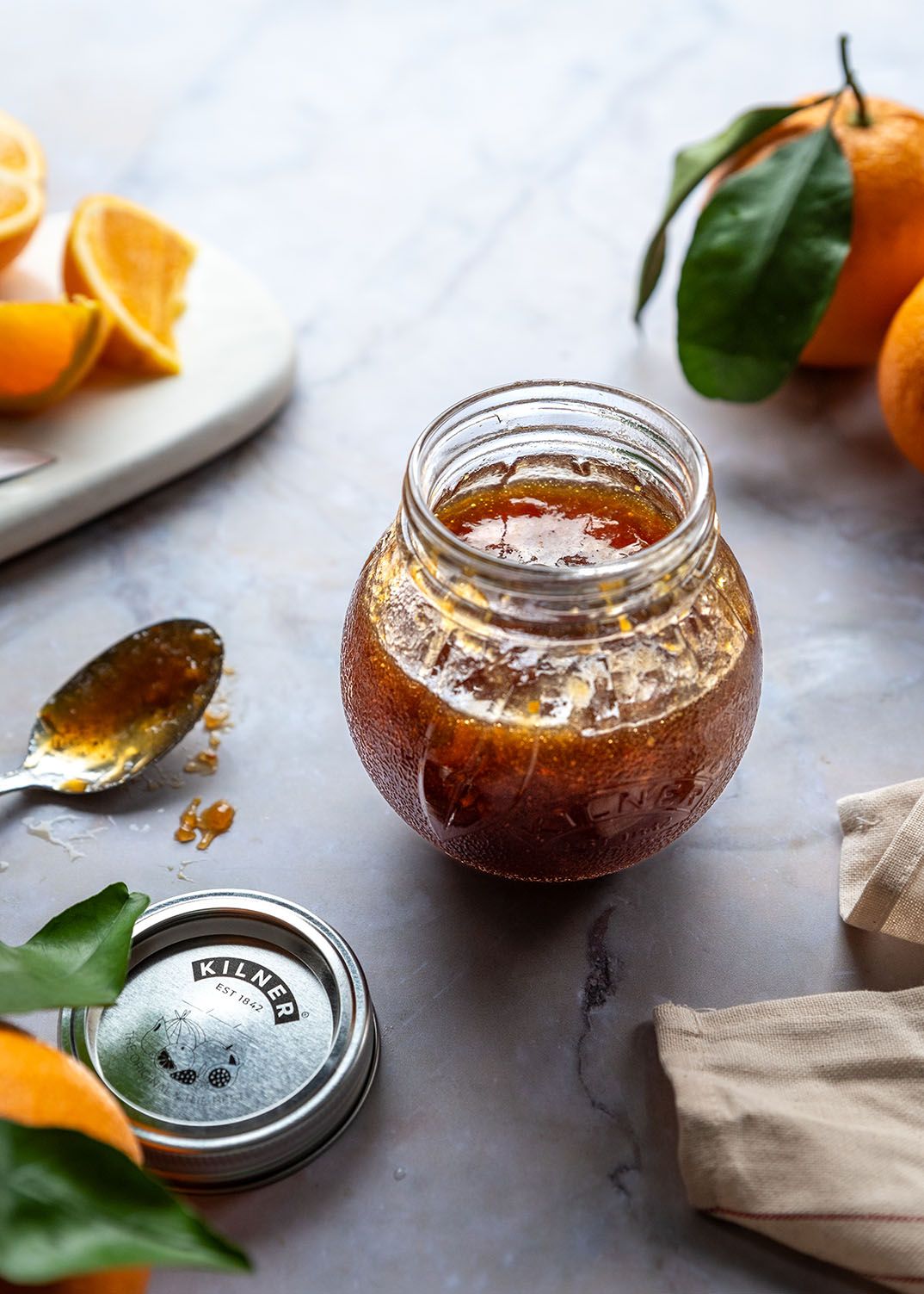 Jar of marmalade with fresh citrus fruits and leaves on marble surface.
