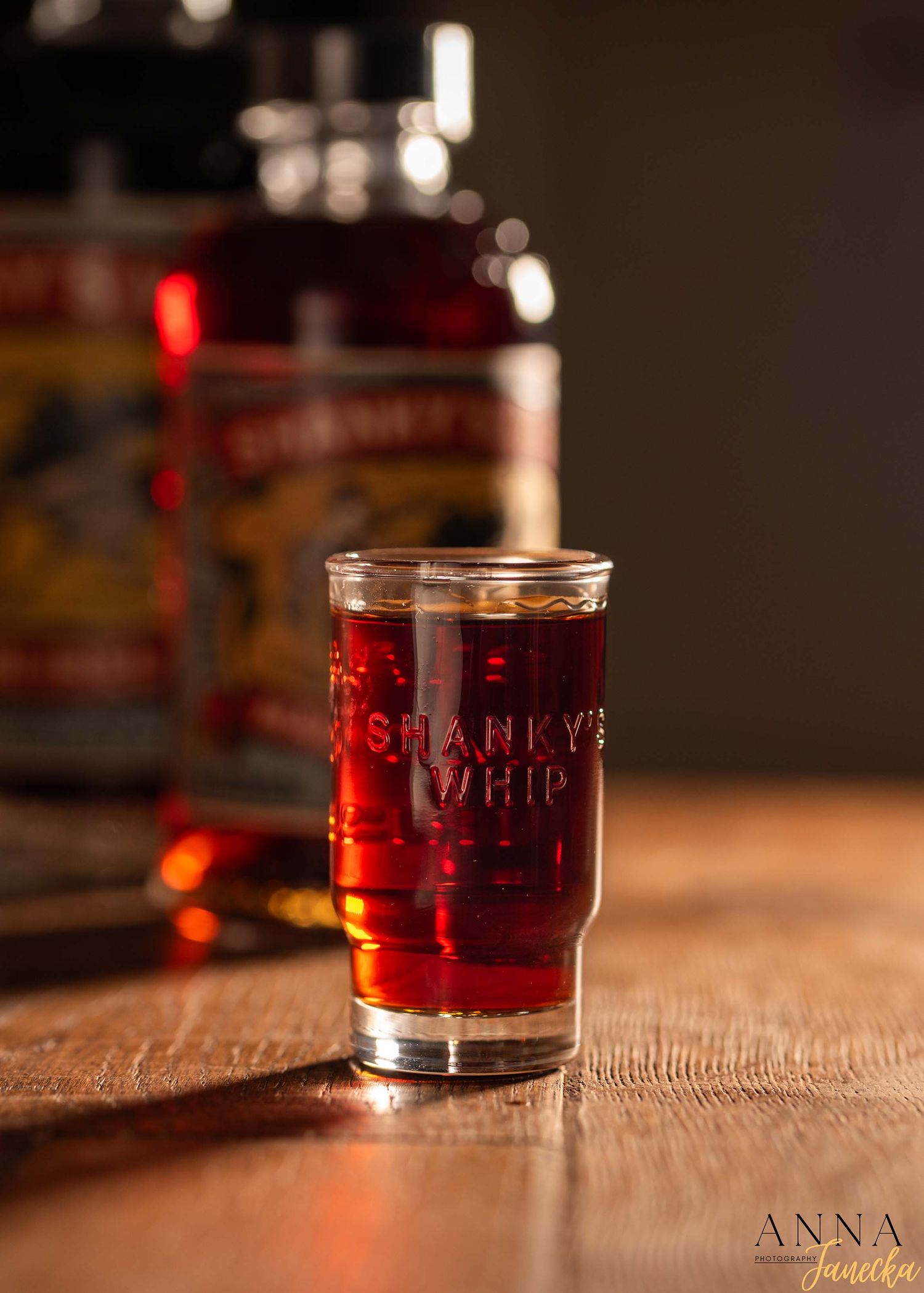 A shot glass filled with red liquor on a rustic wooden table with blurred bottles in background.