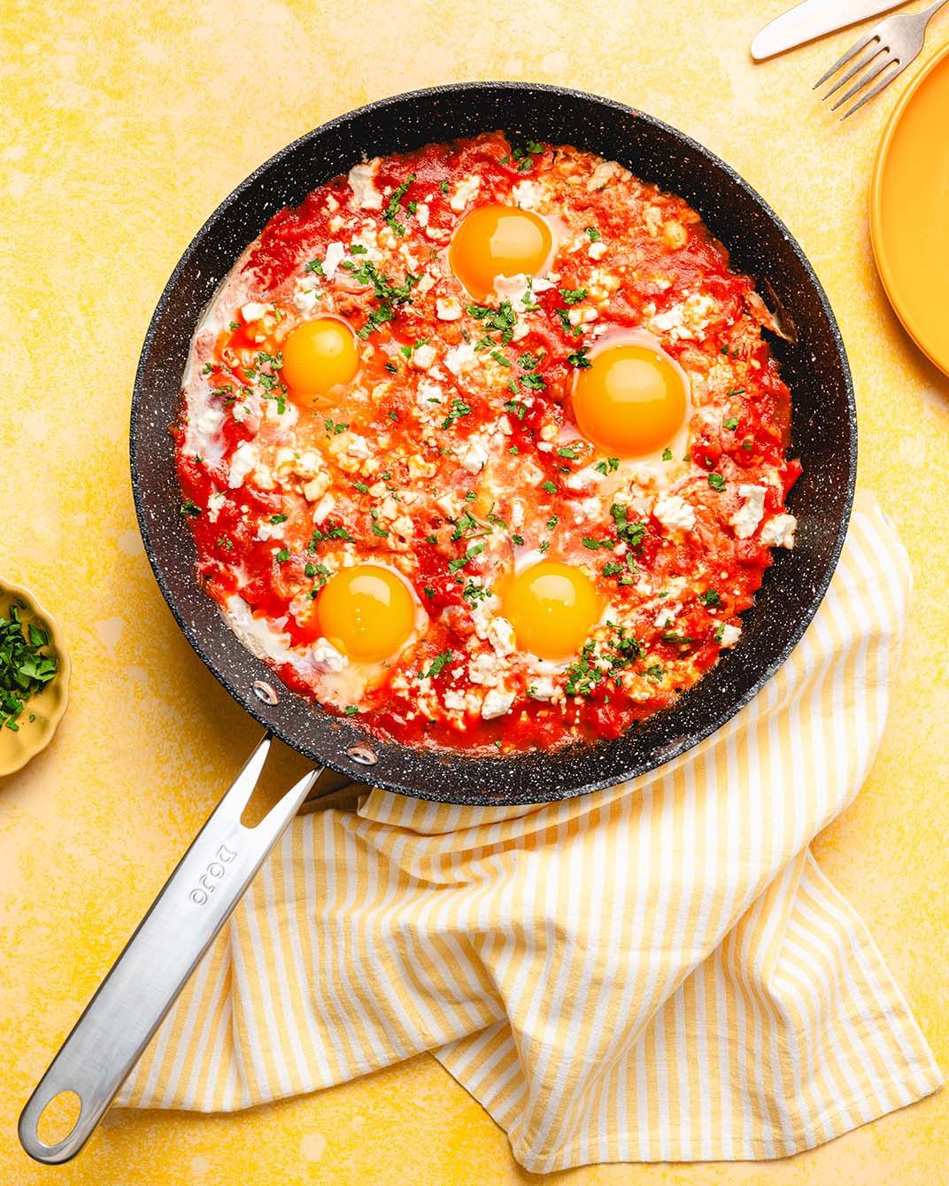 Shakshuka with four eggs poached in spiced tomato sauce, topped with feta and herbs, served in a black skillet.