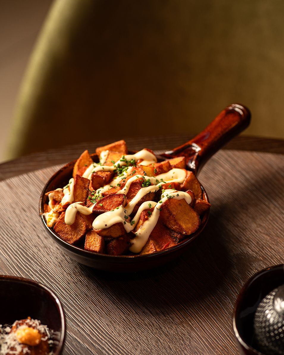 Crispy roasted sweet potatoes topped with creamy white sauce and fresh herbs served in a rustic wooden skillet.