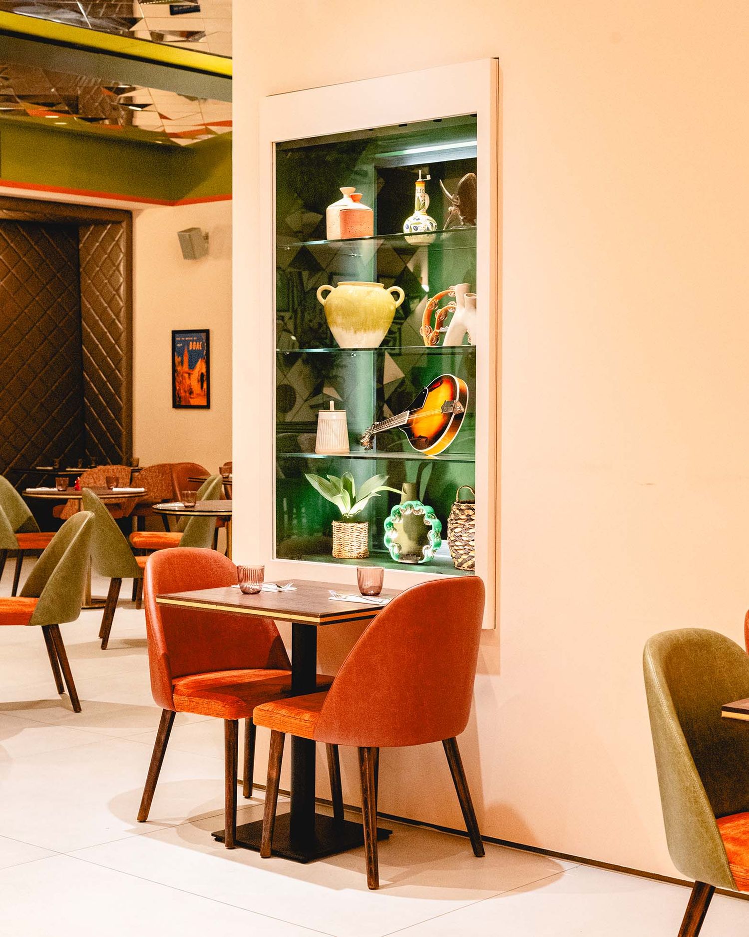 Elegant restaurant interior with orange velvet chairs, wooden table, and green display cabinet showcasing decorative ceramic vases.