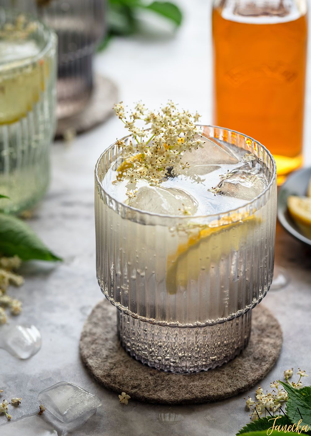 A refreshing cocktail garnished with elderflowers and lemon slice served in a ribbed glass with ice on a stone coaster.
