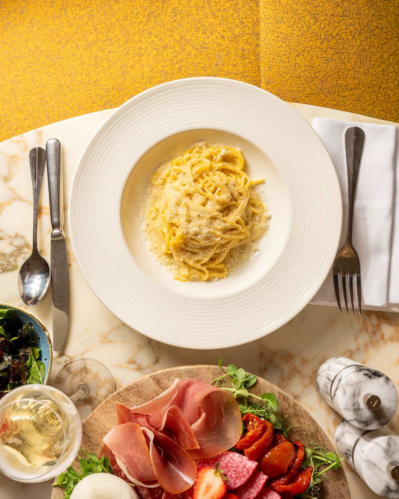 Overhead view of creamy pasta on white plate beside charcuterie board with prosciutto, salami, and fresh strawberries.
