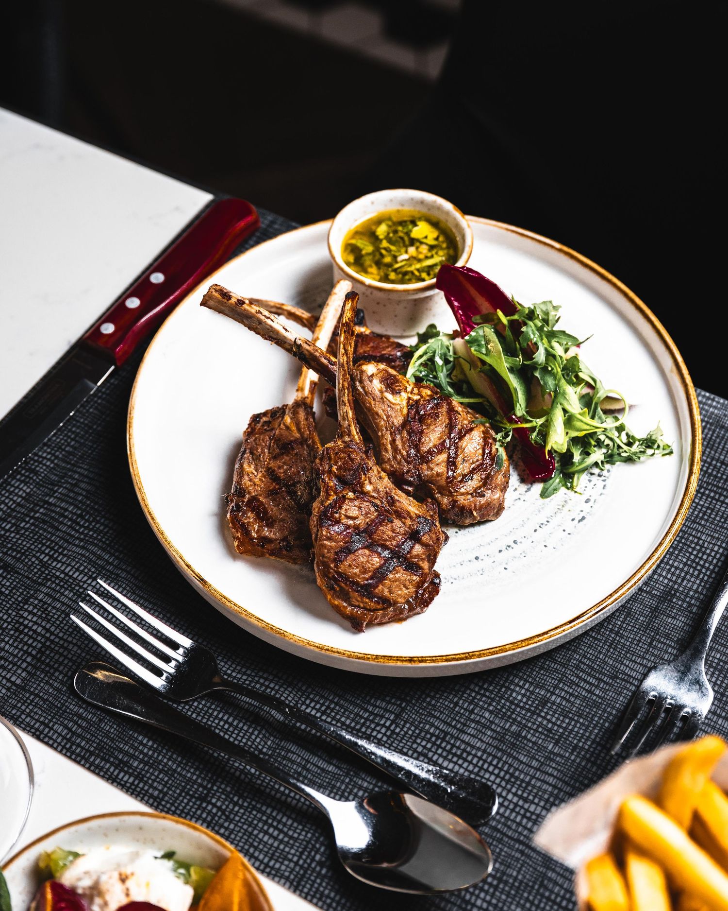 Grilled lamb chops on a white plate with fresh greens, beet, and a small bowl of herb sauce at a restaurant.